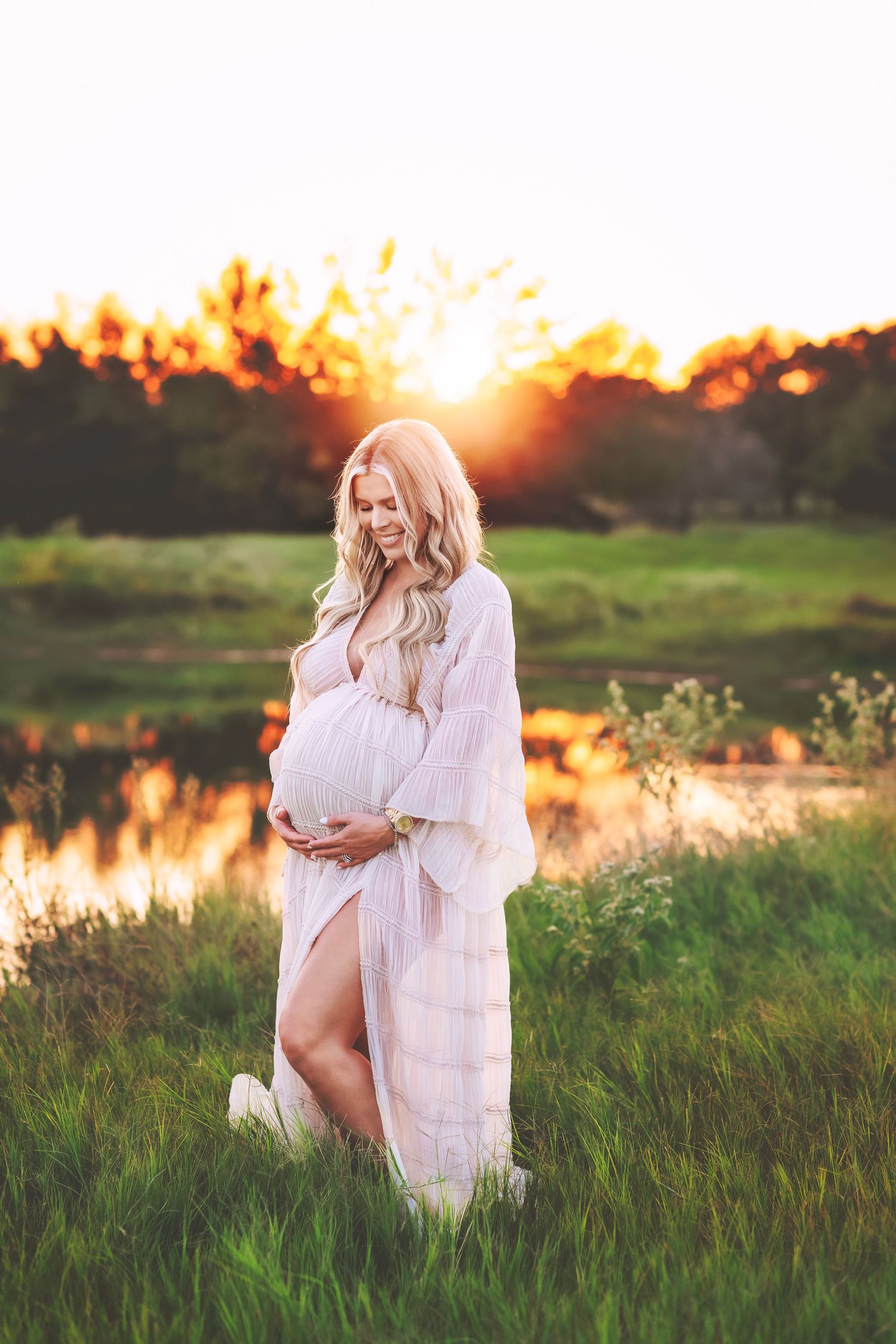 Ethereal maternity portrait of woman in white dress during golden sunset by lake.