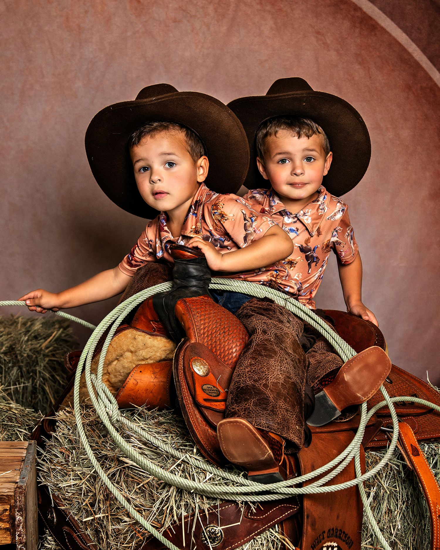 Two young cowboys in matching outfits sitting on hay bales with western props.
