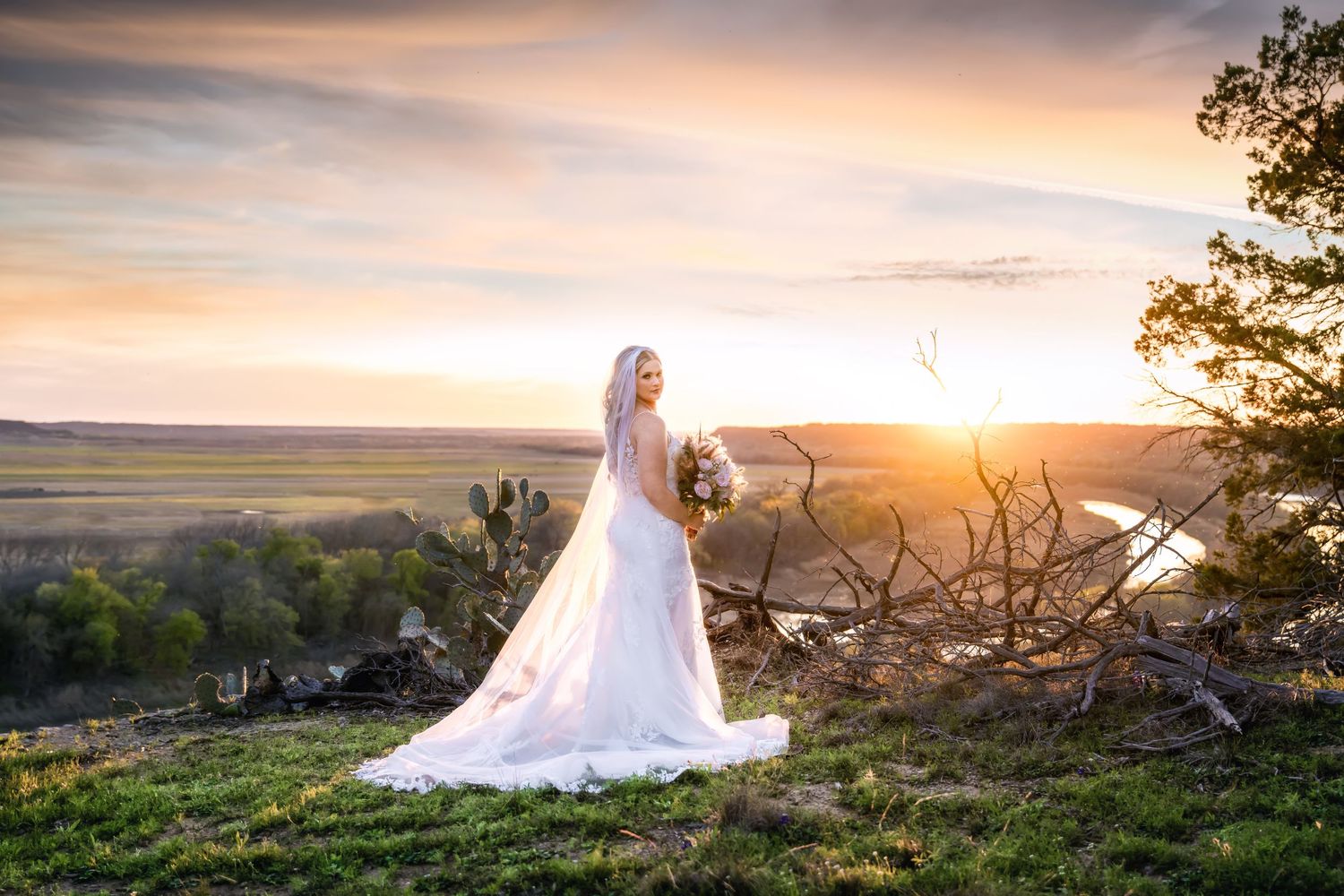 A bride in a flowing veil stands on a hilltop during a golden sunset.