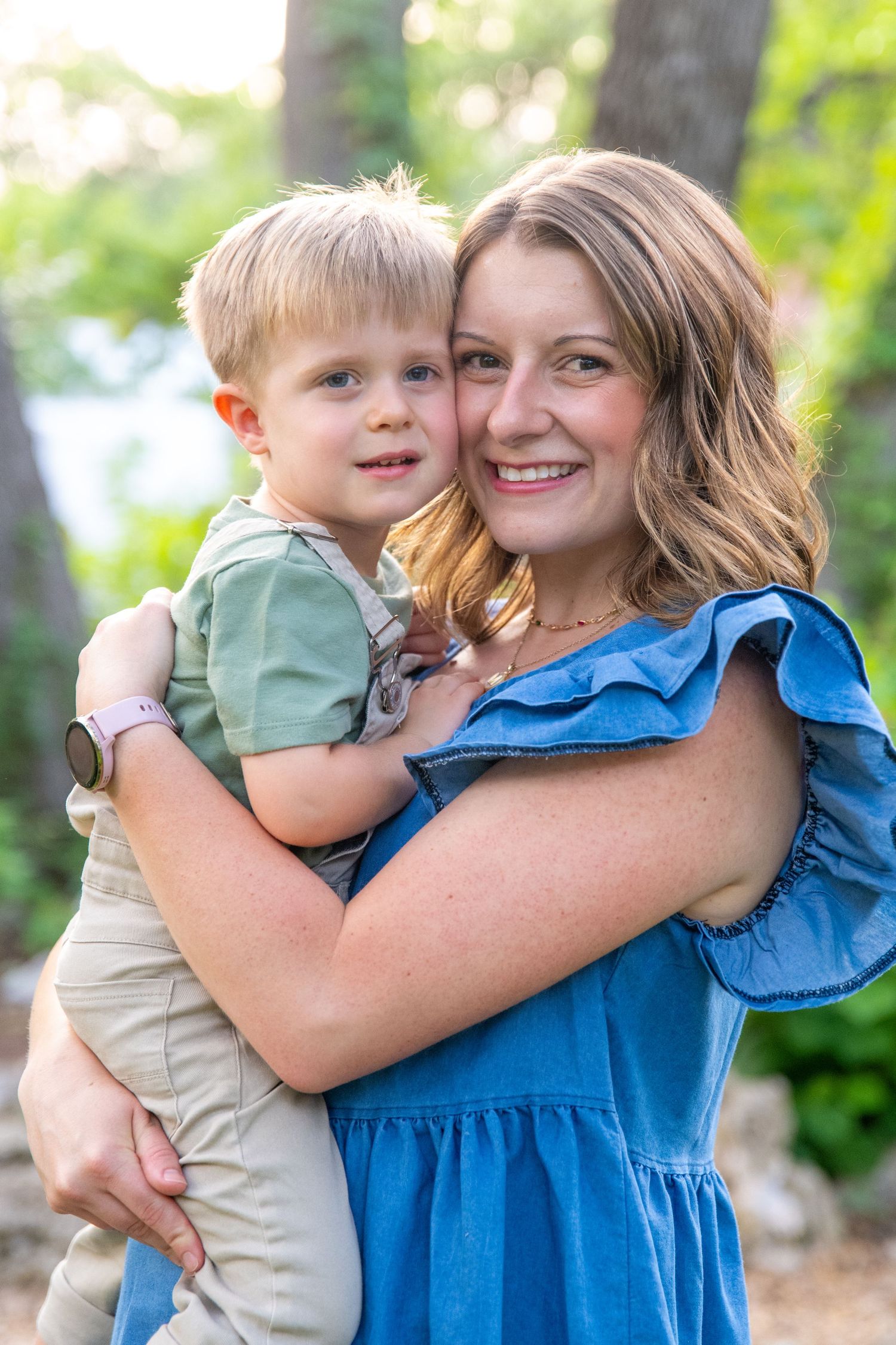 Mother in blue dress holding young child while posing outdoors in natural setting.