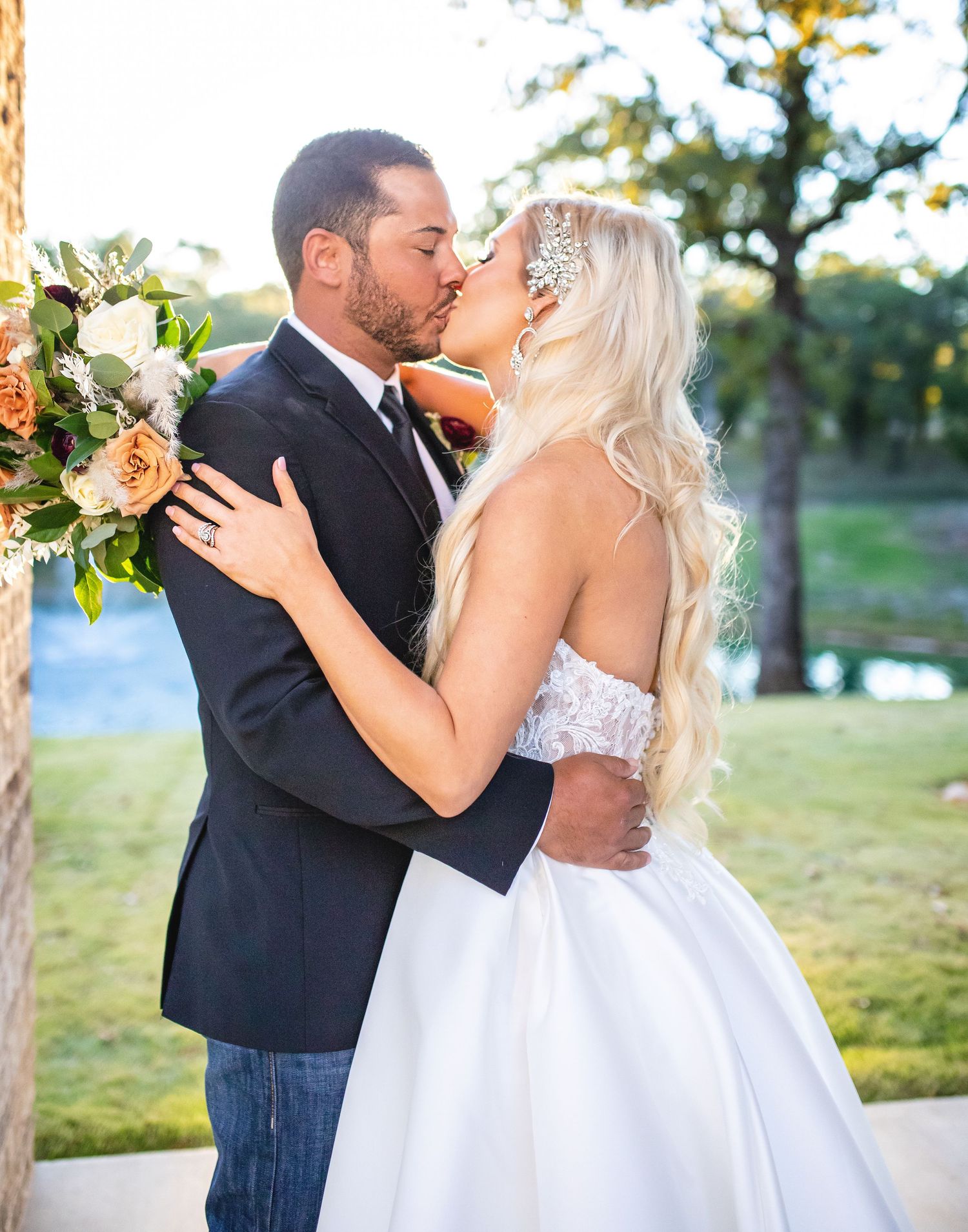 A wedding couple shares a romantic kiss outdoors with the bride holding a peach and white floral bouquet.