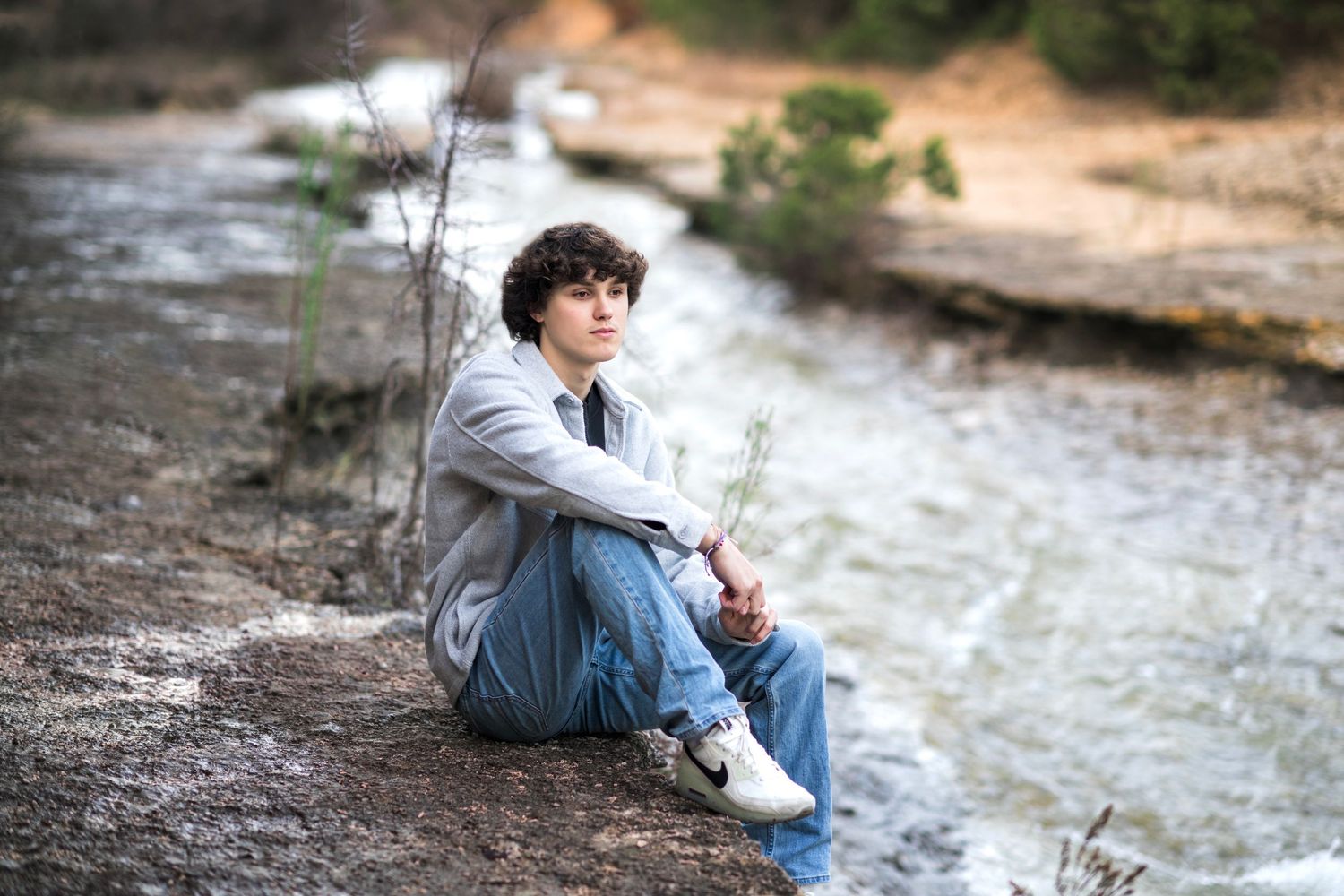 Person in casual attire sits thoughtfully by waterfront on rocky shore.