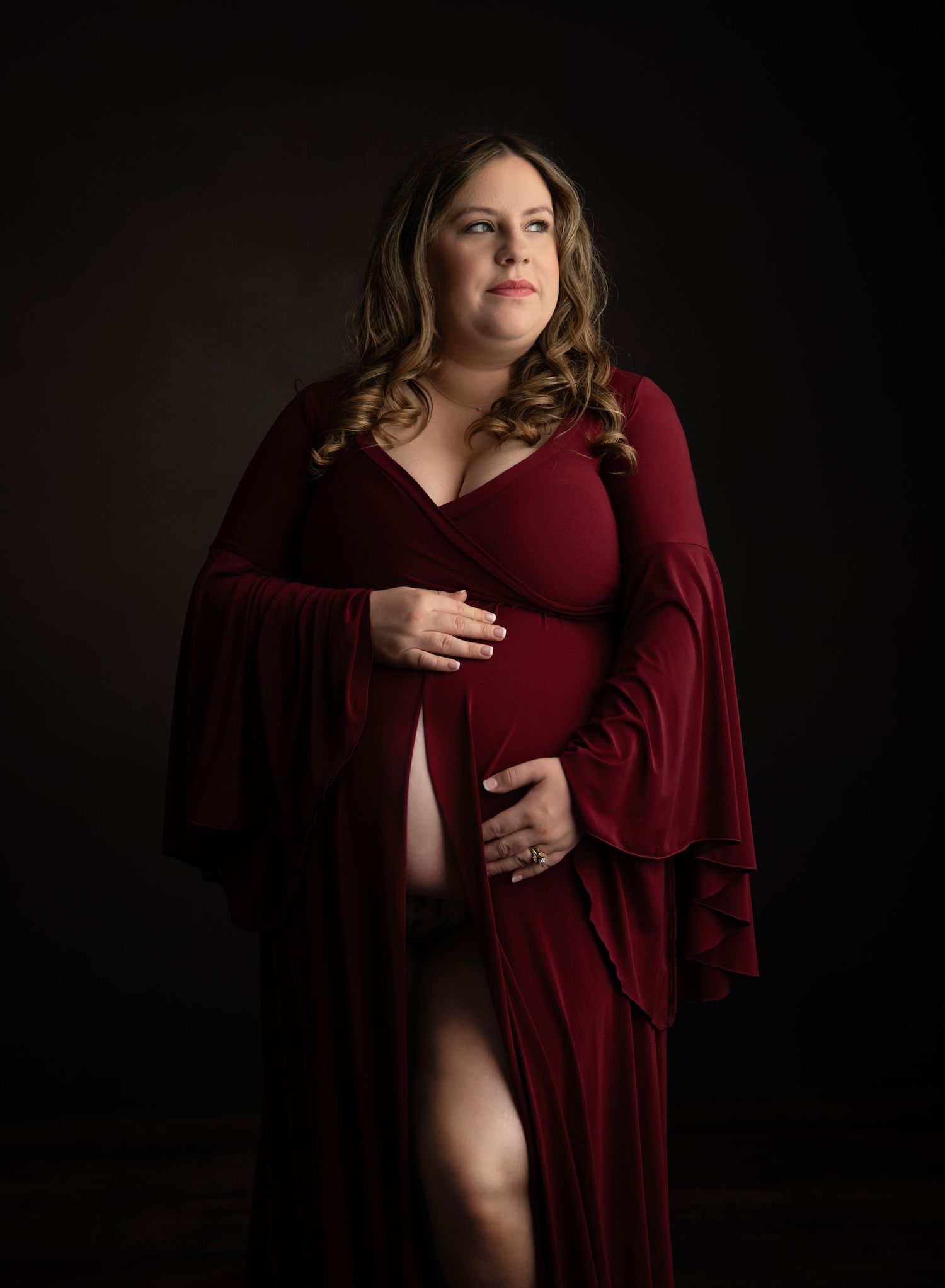 Dramatic maternity portrait with woman in burgundy velvet dress against dark background.