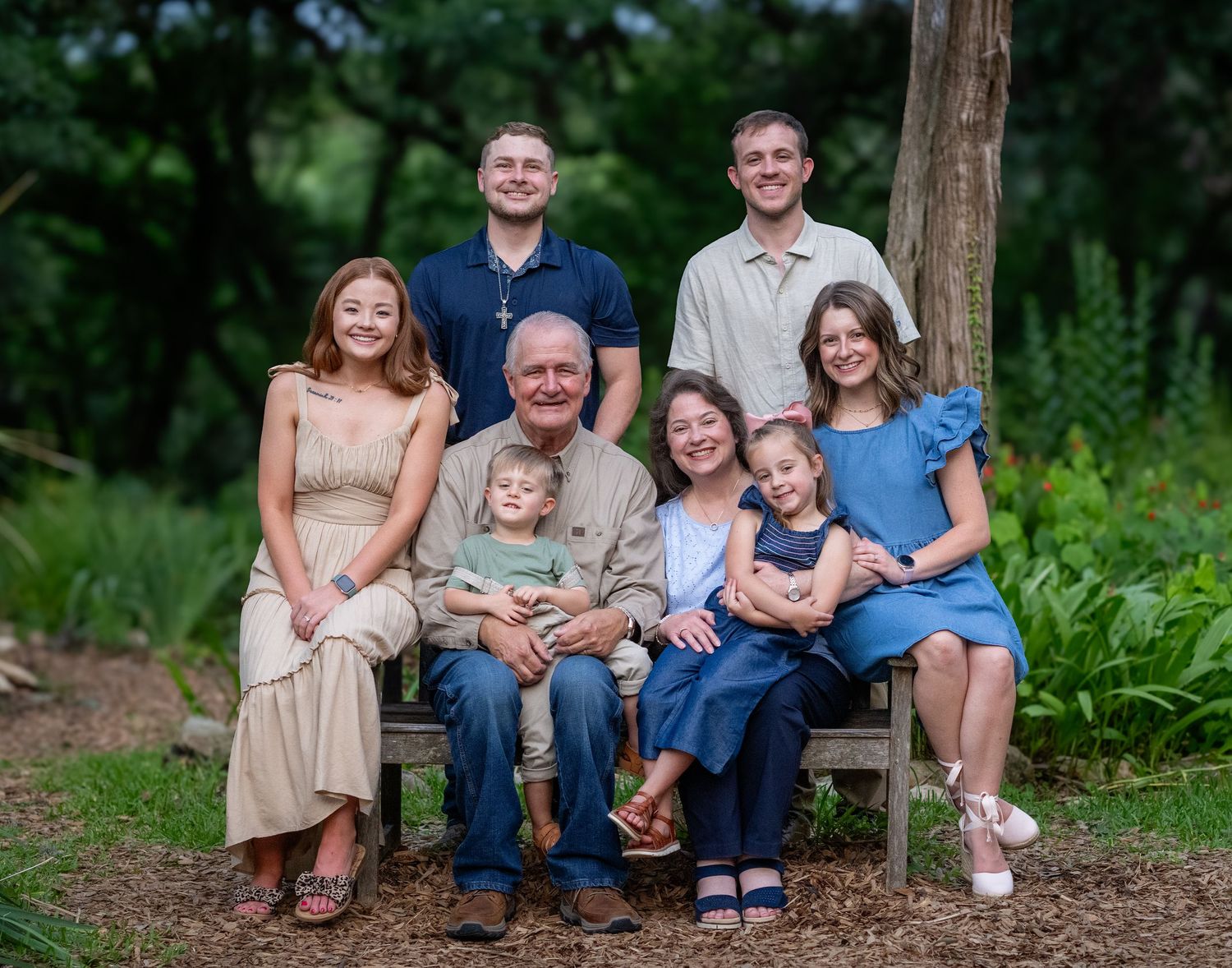 Multi-generational family poses together on a wooden bench in a lush garden setting.