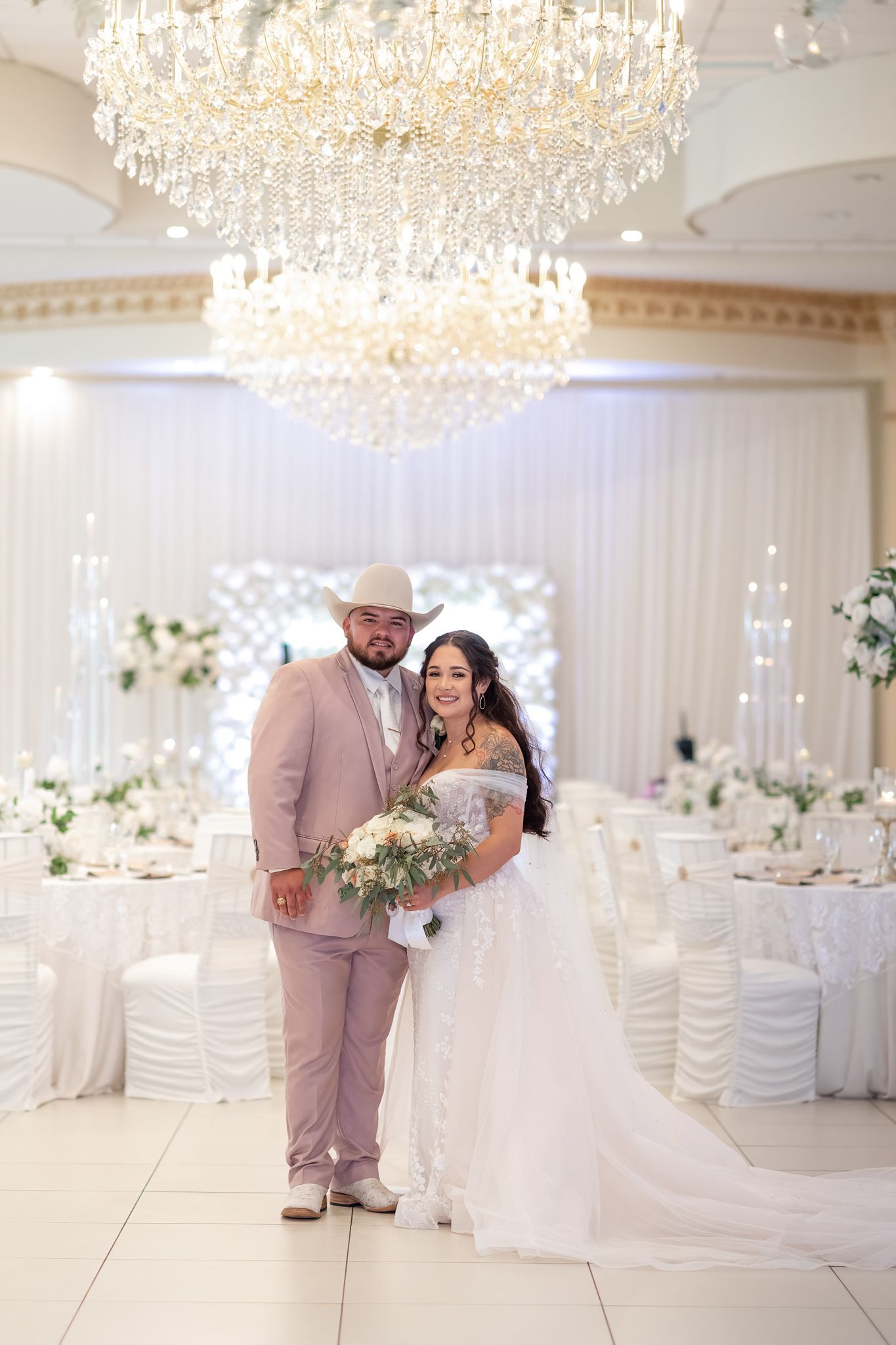 Elegant wedding portrait under crystal chandelier in white decorated ballroom.