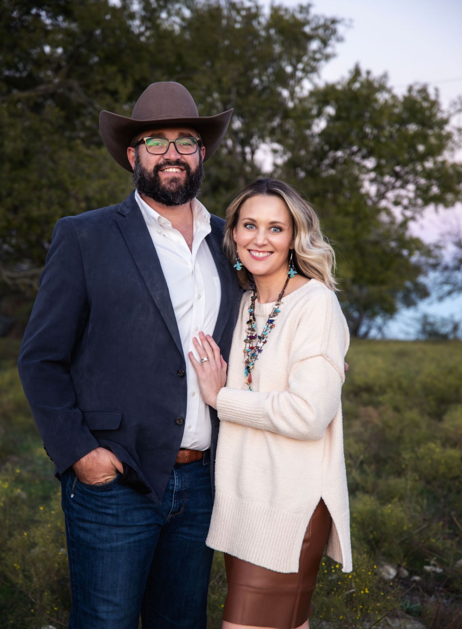 Couple in western attire poses outdoors during a rustic engagement photoshoot.