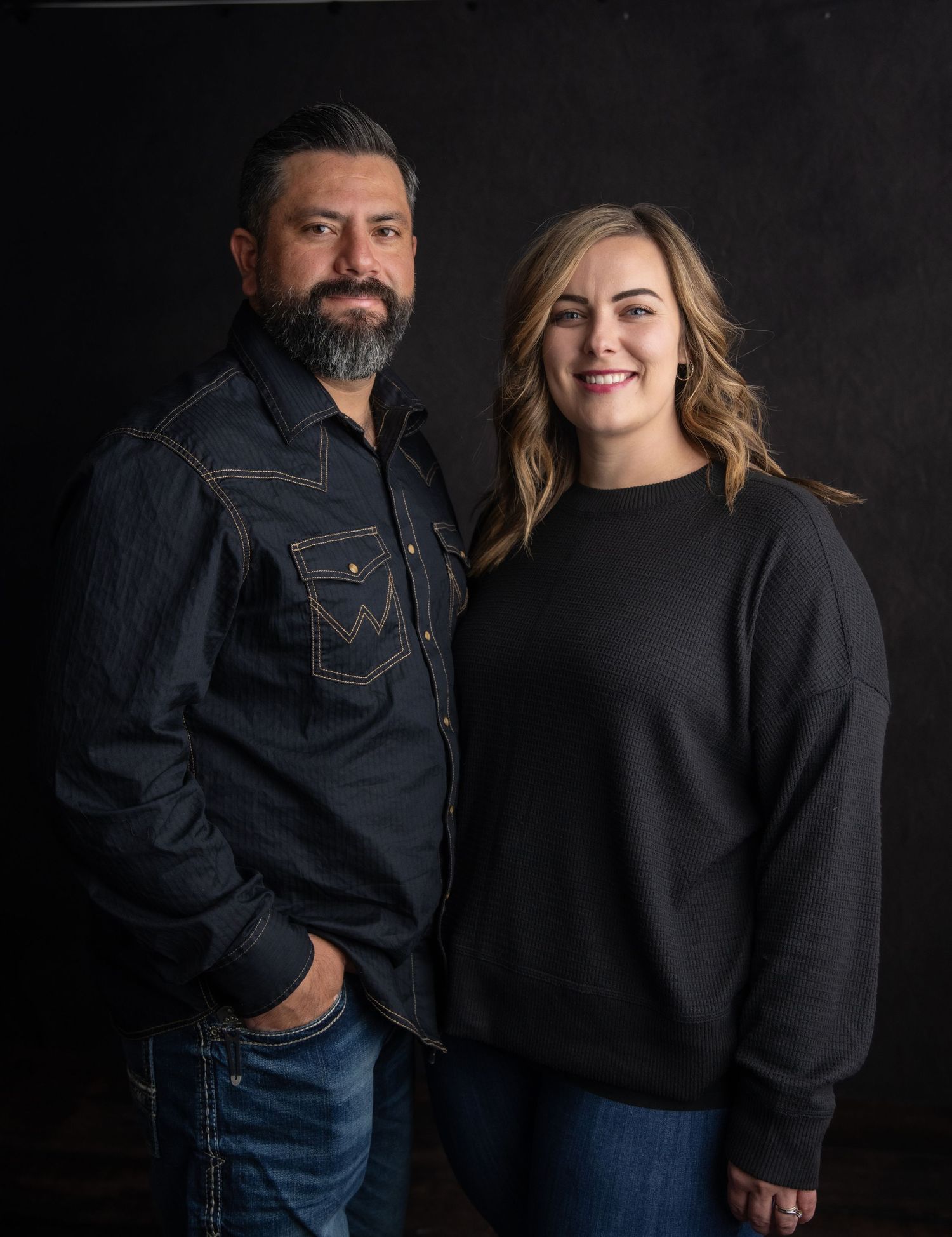 Two people in casual attire pose together against a dark background in a studio portrait setting.