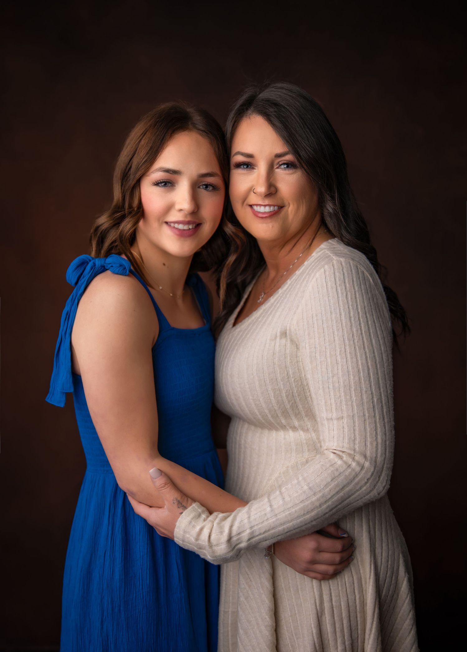 Portrait of two people in a studio setting wearing a royal blue dress and cream sweater against a dark brown background.