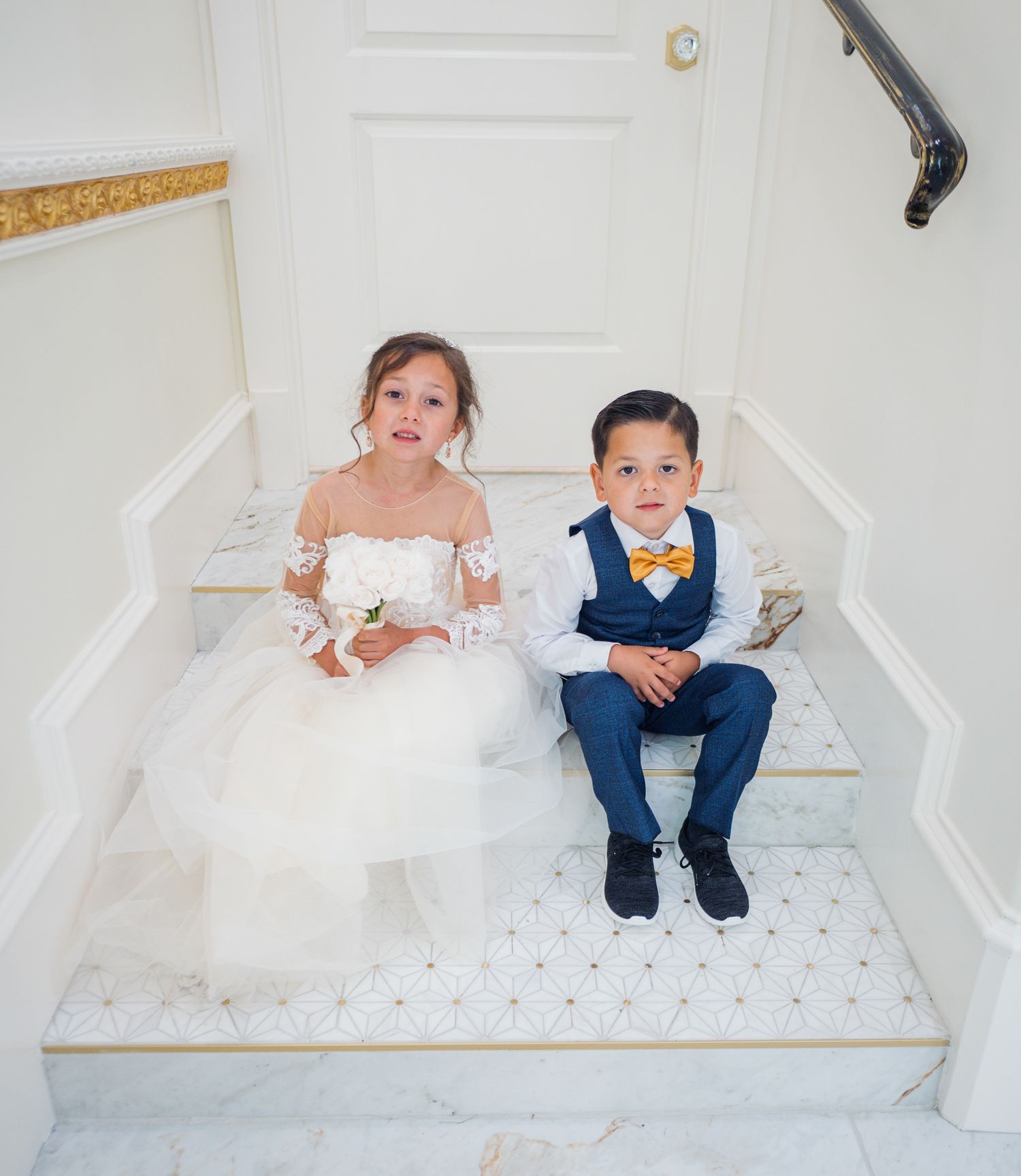 Two children in formal wear sitting on white marble stairs.