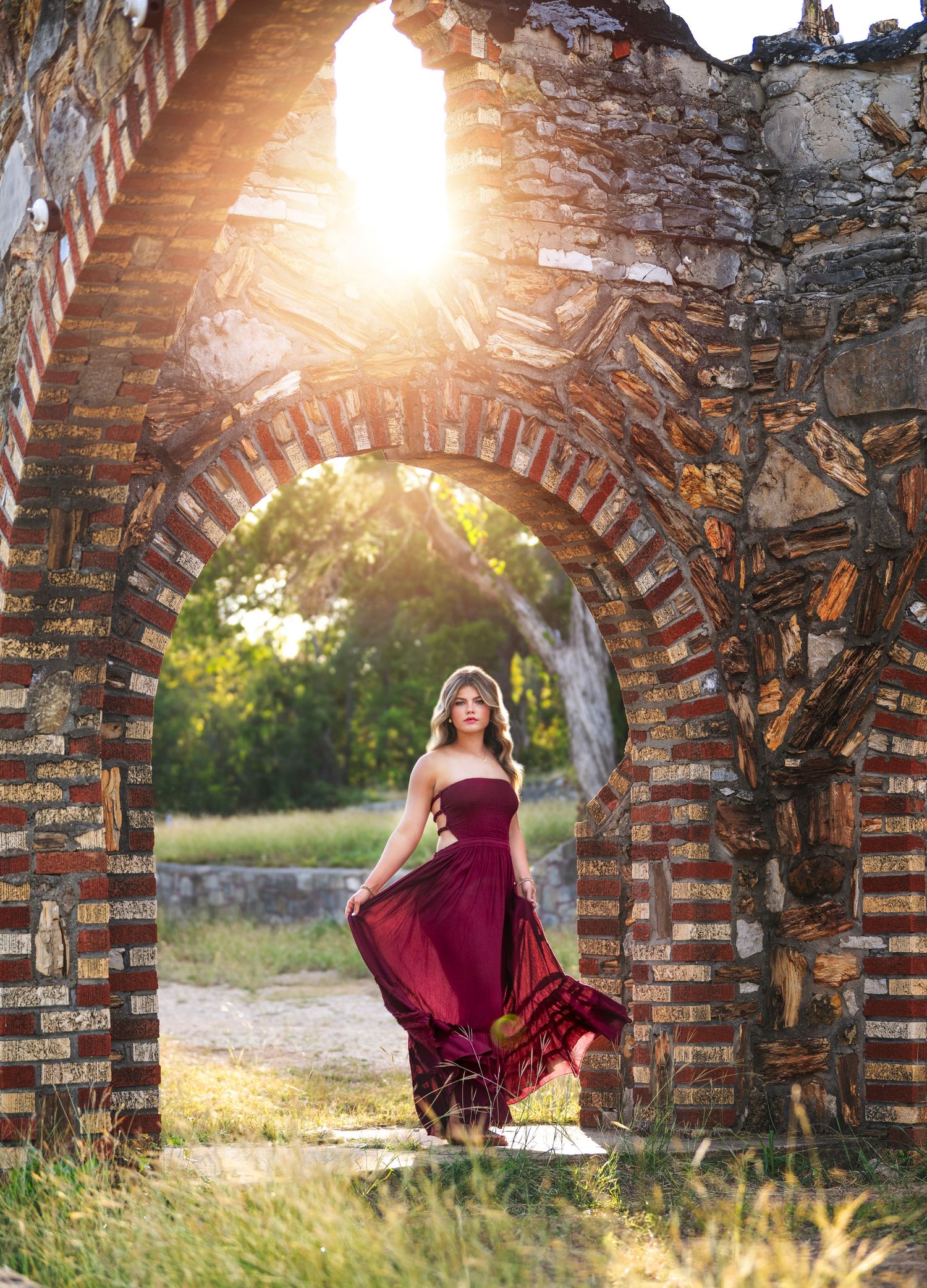 A flowing burgundy dress twirls gracefully through stone archways at sunset in an outdoor setting.
