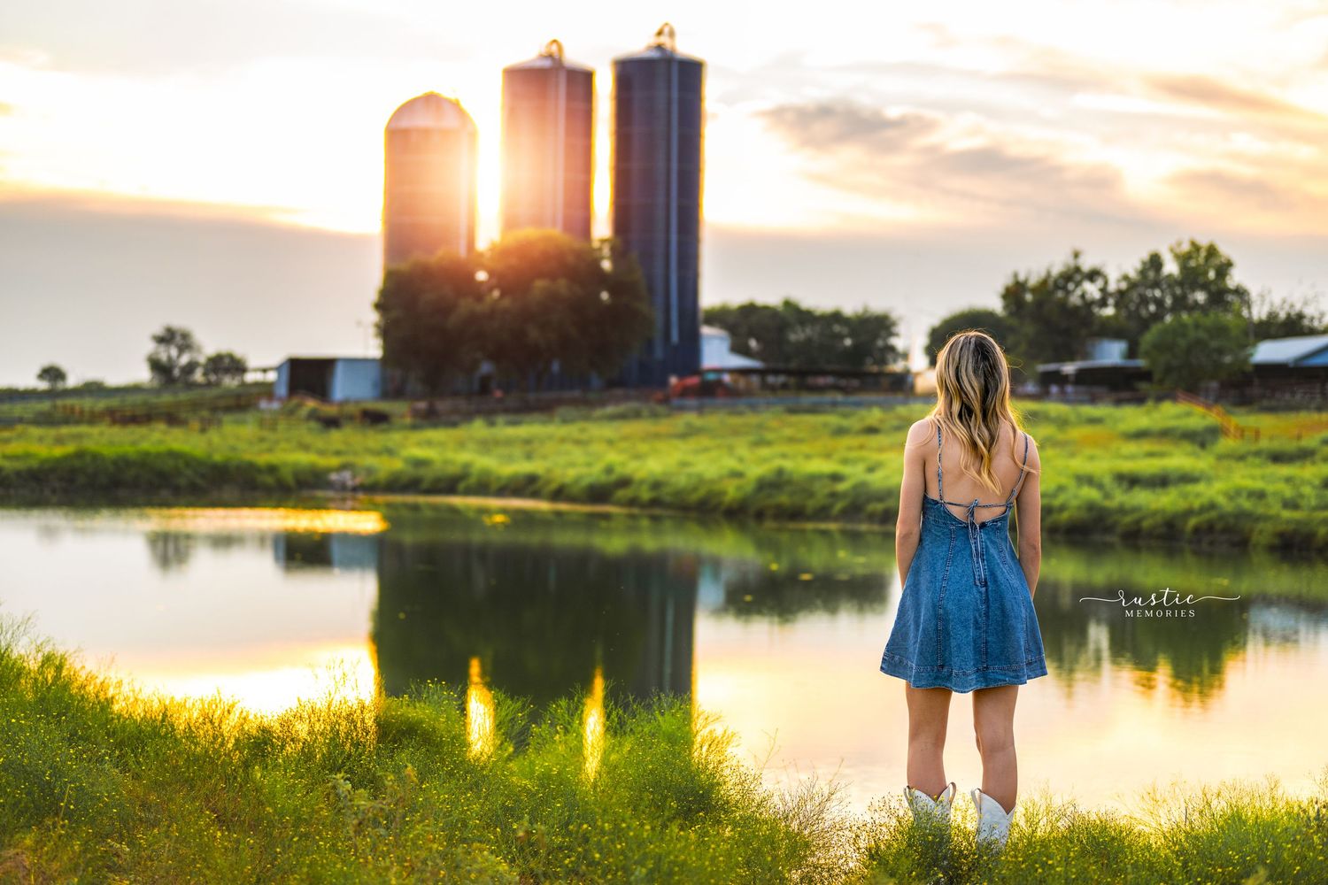 A couple stands together at sunset near a serene pond with farm silos in the background creating a romantic rural scene.