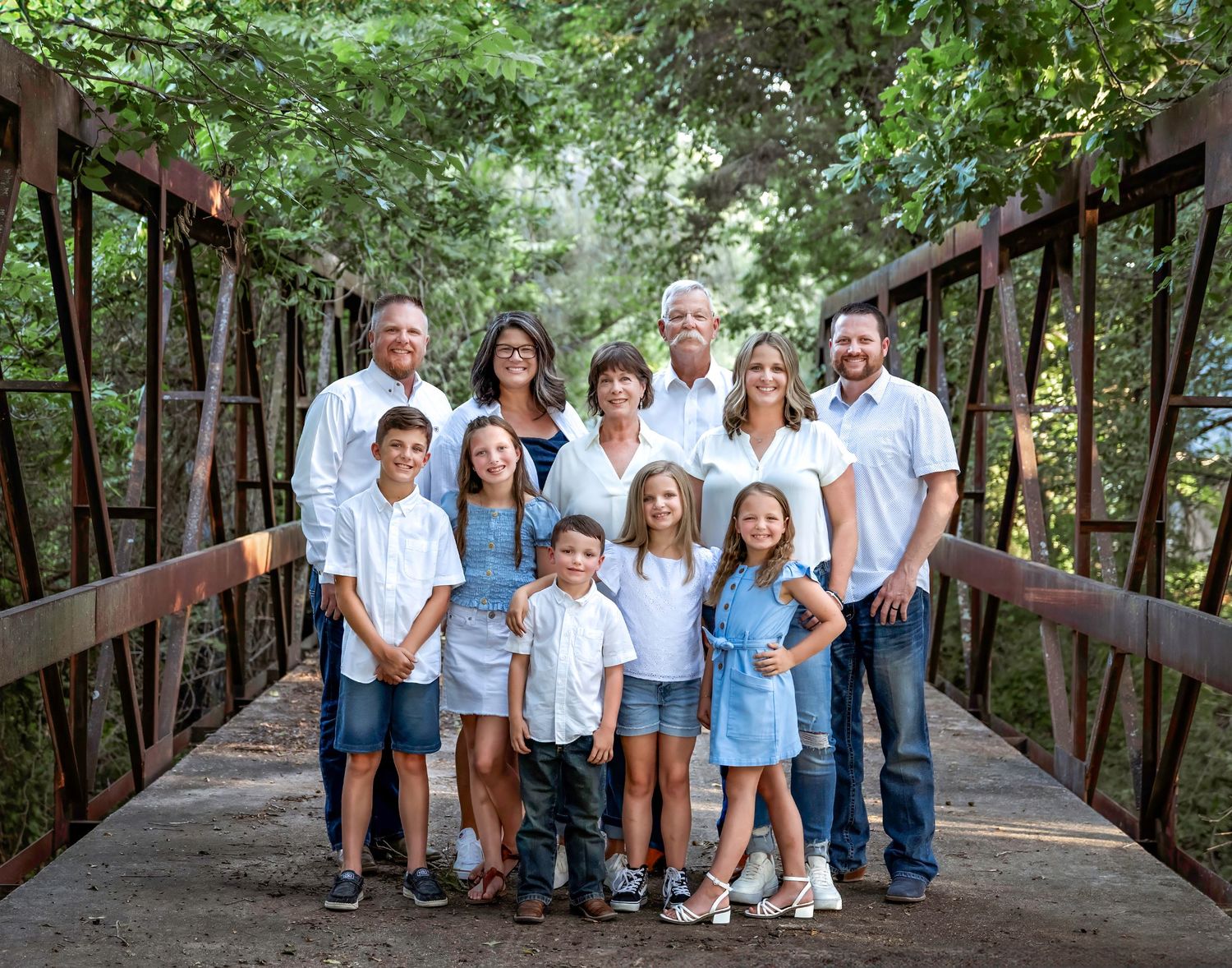 A multi-generational family poses on a wooden bridge surrounded by lush green trees.