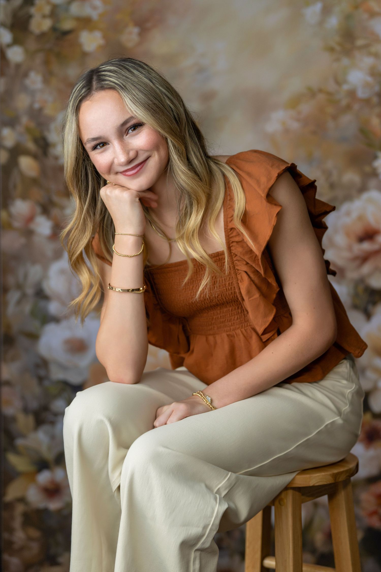 Person in brown sleeveless top and white pants sits casually on wooden stool against autumn-toned background.