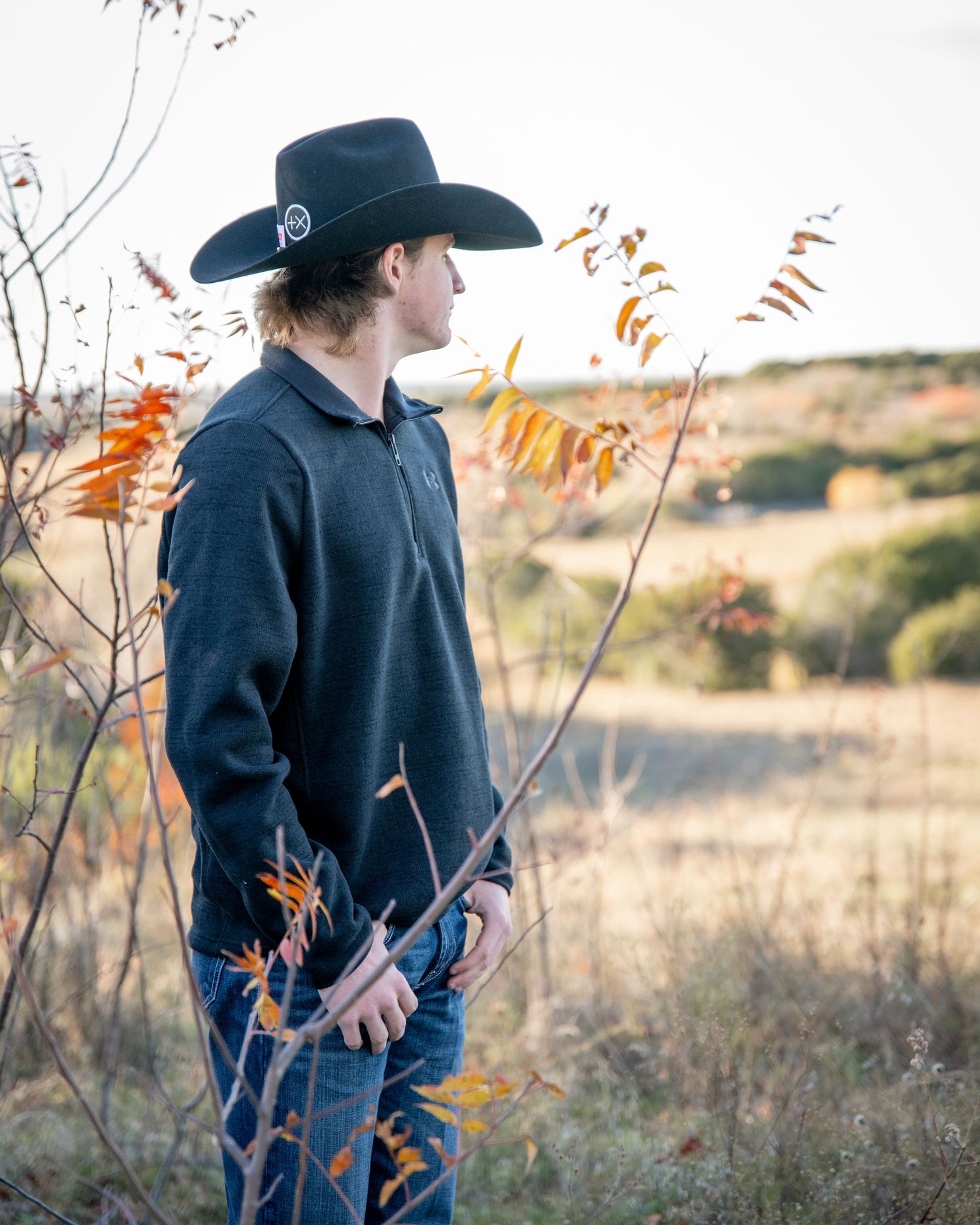Figure in dark cowboy hat and navy sweater stands among autumn foliage at sunset.