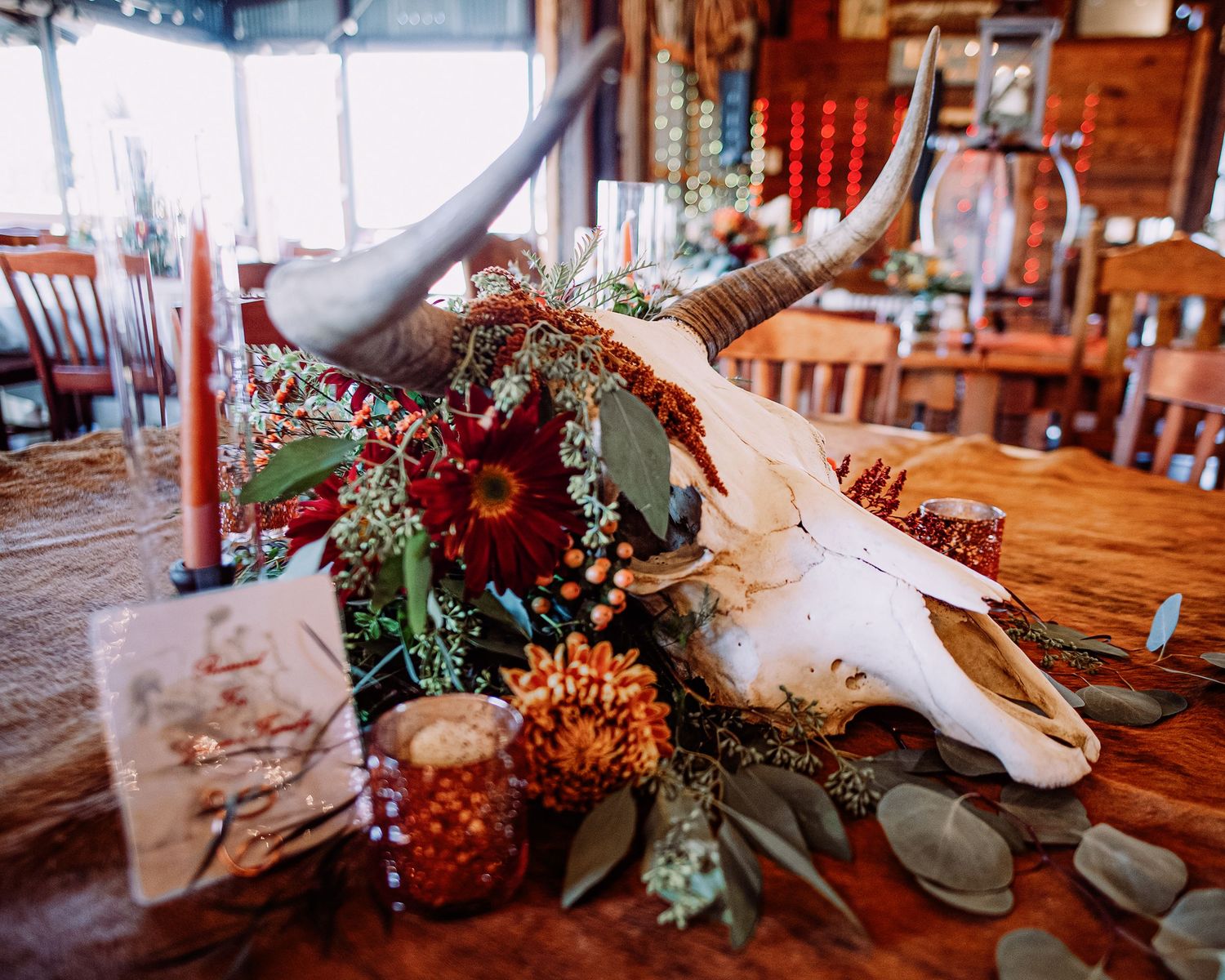 Rustic centerpiece featuring animal skull, dark red flowers, and greenery on wooden table.