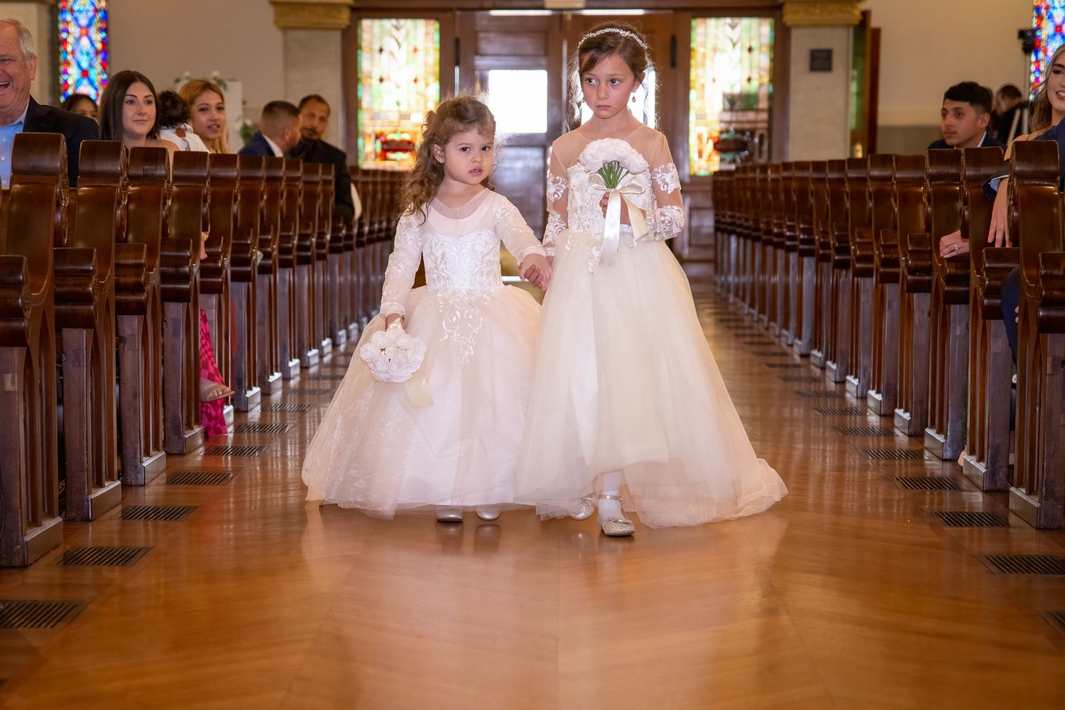 Two flower girls in white dresses walking down the aisle of a church during a wedding ceremony.