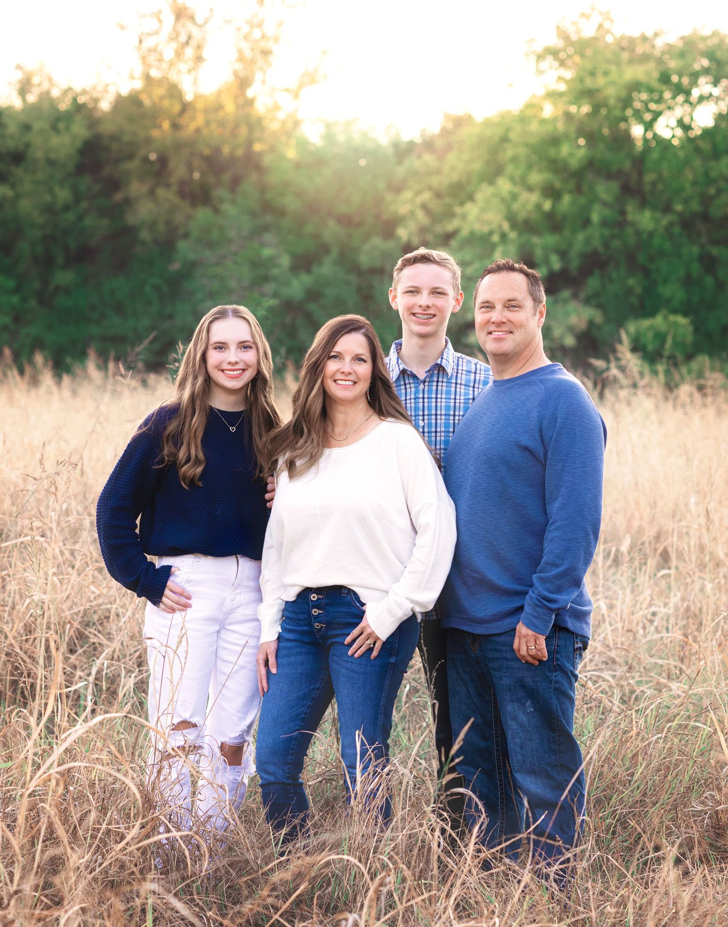 A family of four poses in casual attire against a sunset backdrop in tall grass.