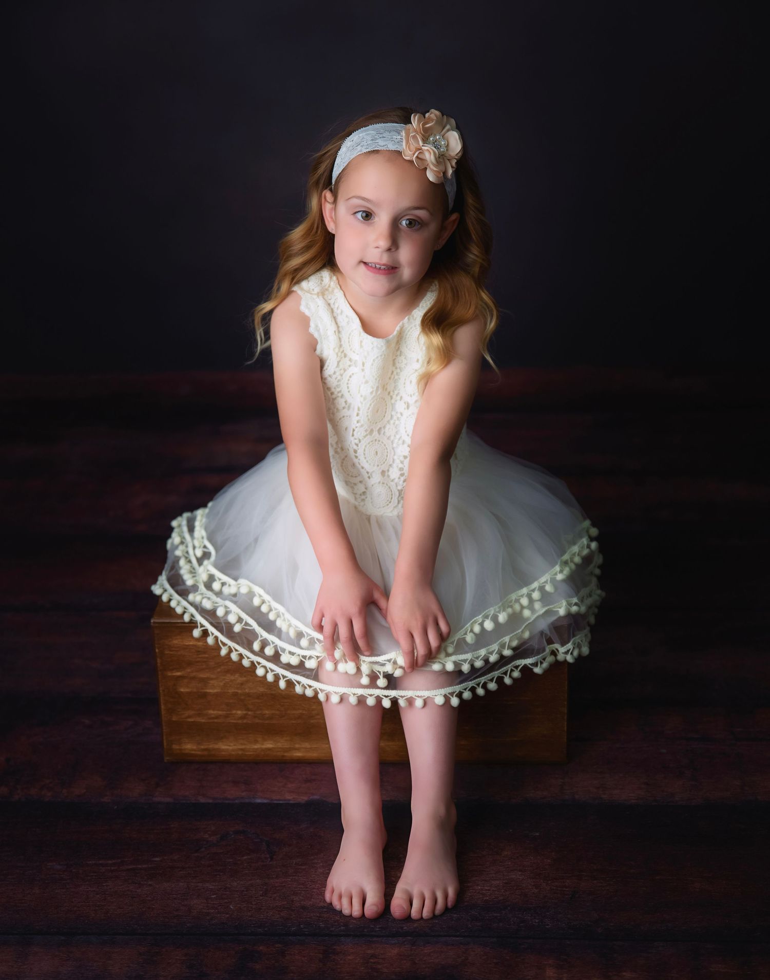 Child in white lace dress sits thoughtfully on wooden bench during portrait session.