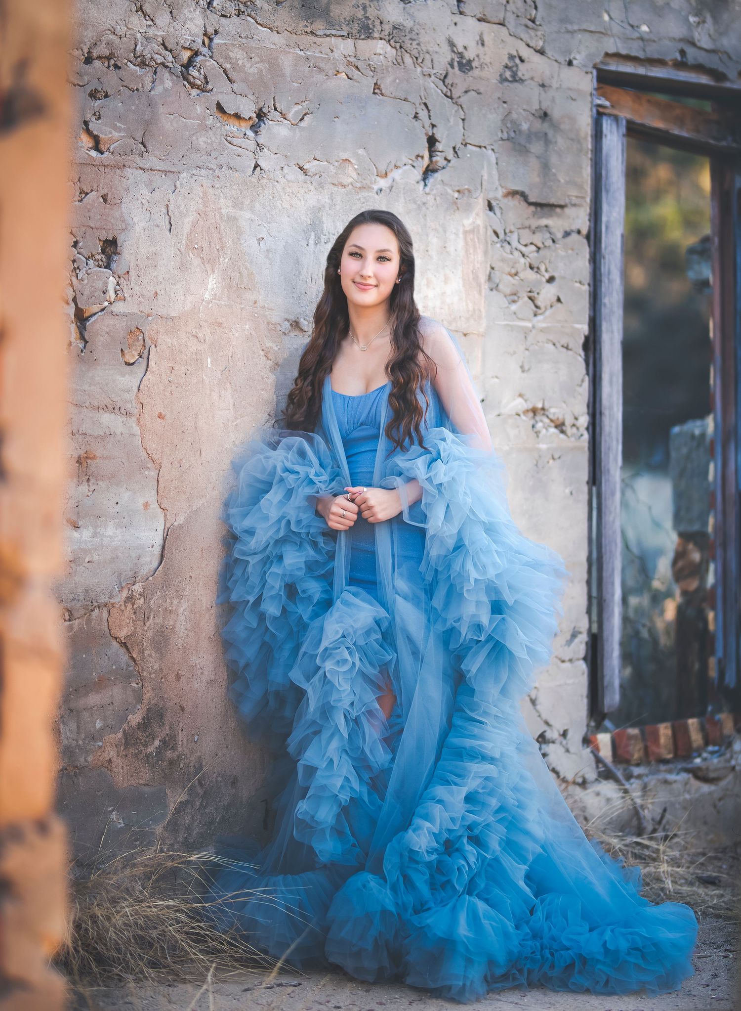 Person in flowing blue gown poses against rustic stone wall.