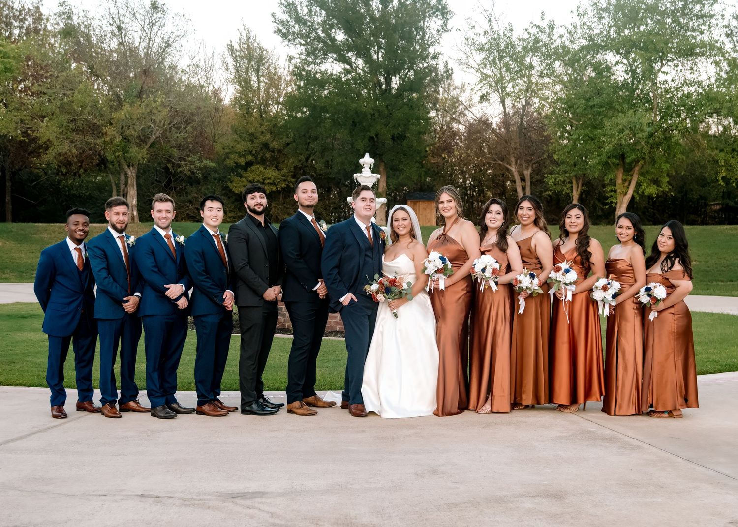 Wedding party in blue suits and rust colored dresses posing together on grass.