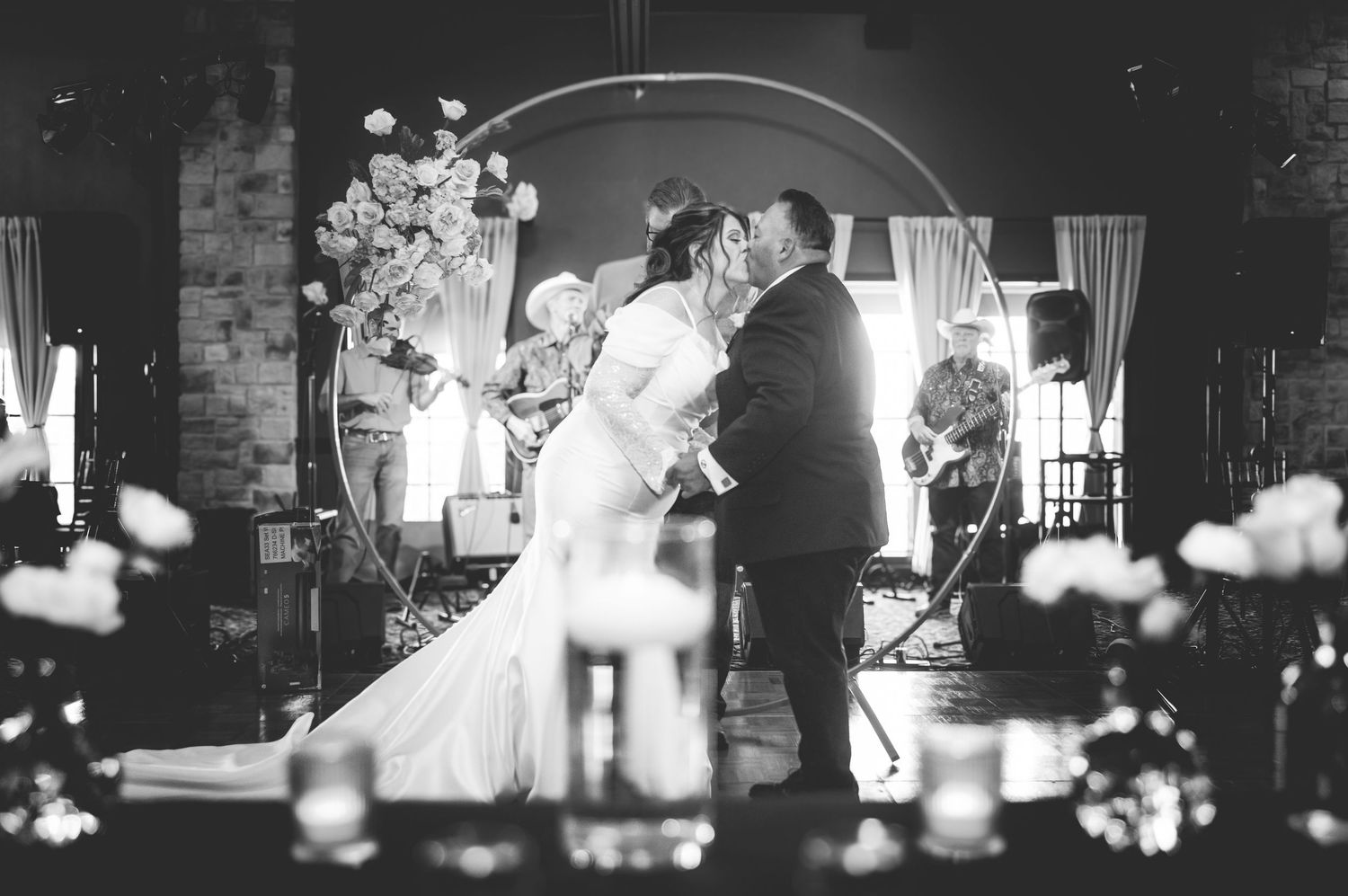 Black and white wedding ceremony photo capturing a couple's first kiss under decorative lighting.