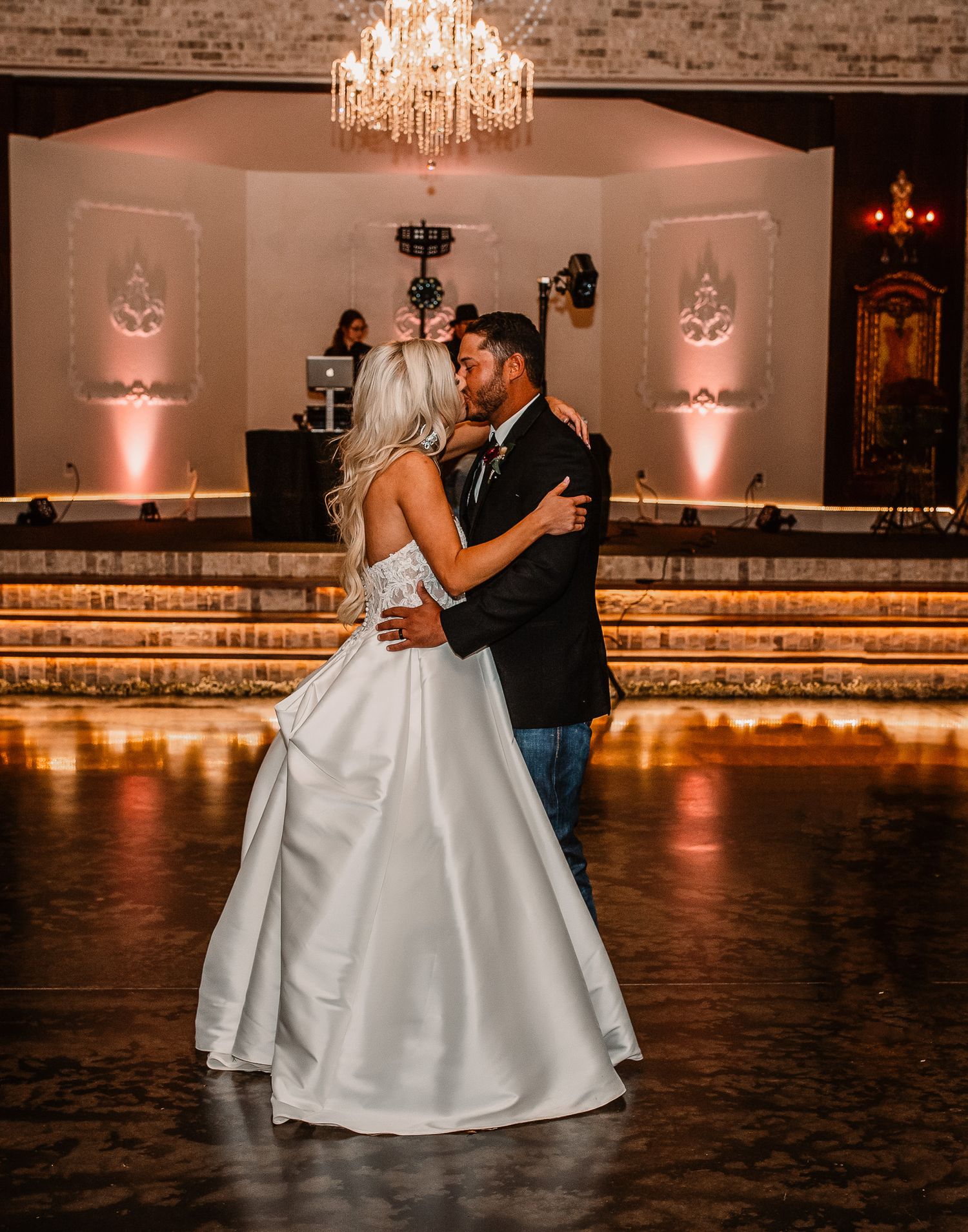 Couple dancing in formal attire under crystal chandelier on dark wooden dance floor.