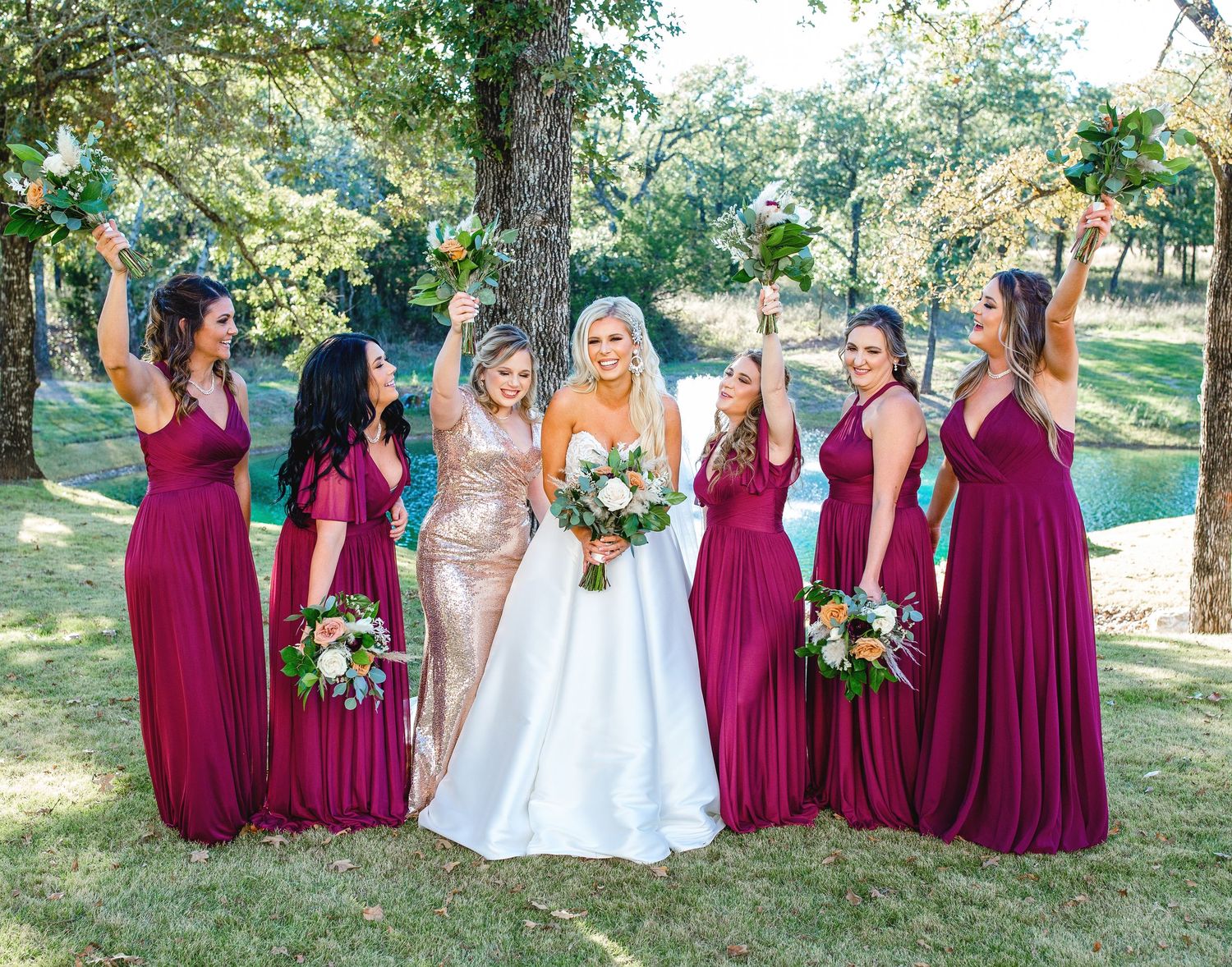 Bridesmaids in burgundy dresses pose with bride in white gown outdoors.