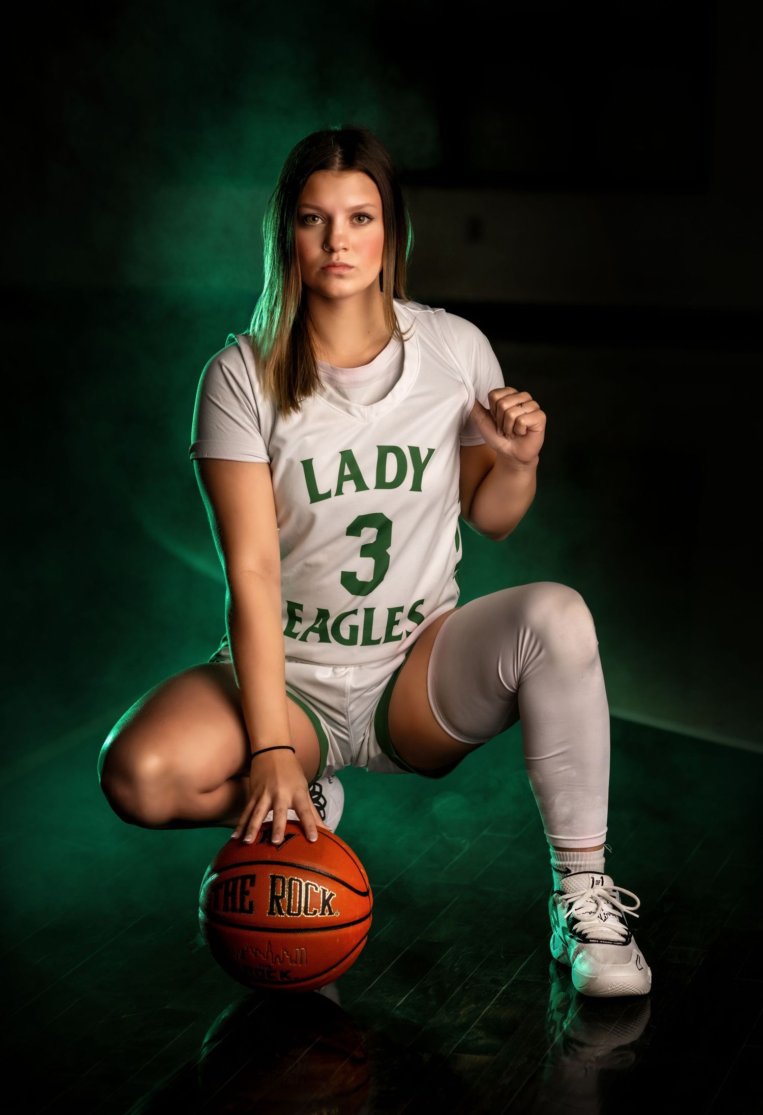 Basketball player in white uniform posing with ball against dark background with green lighting.
