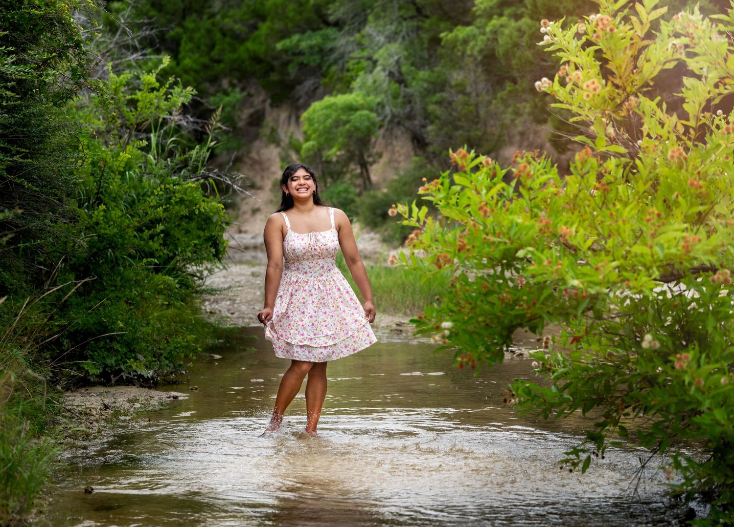 Woman in white dress wades through a flowing stream surrounded by lush greenery.