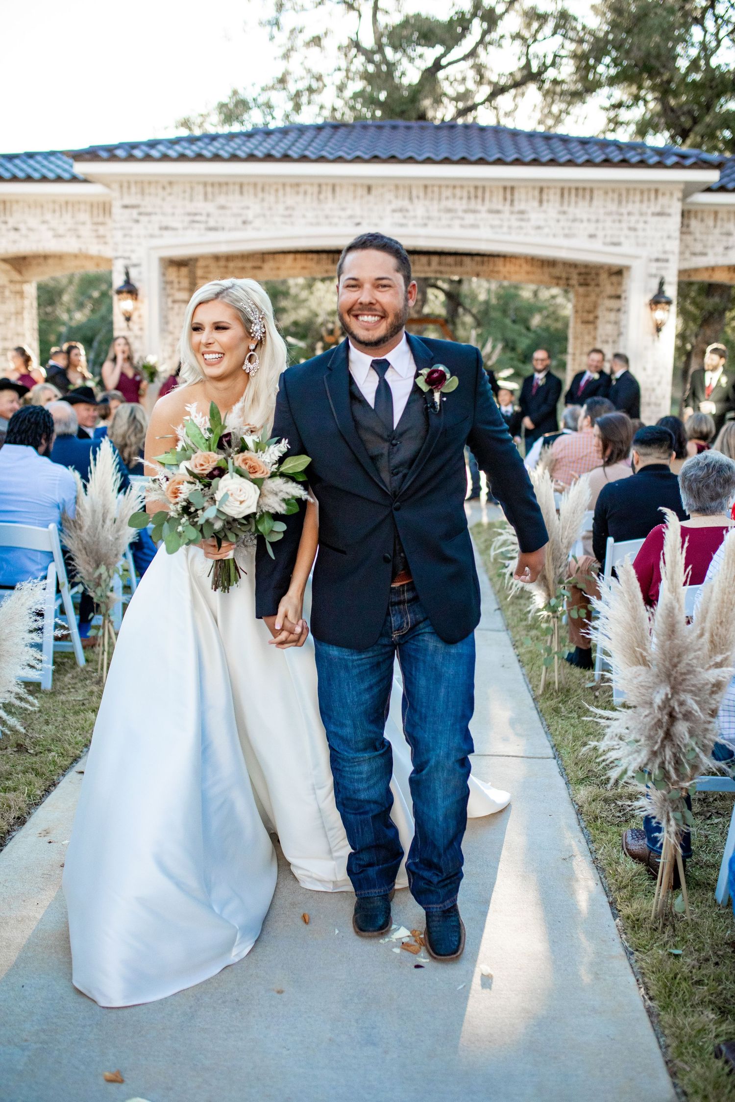 A couple walks down an outdoor aisle after their wedding ceremony at a venue with a stone archway and rustic decor.