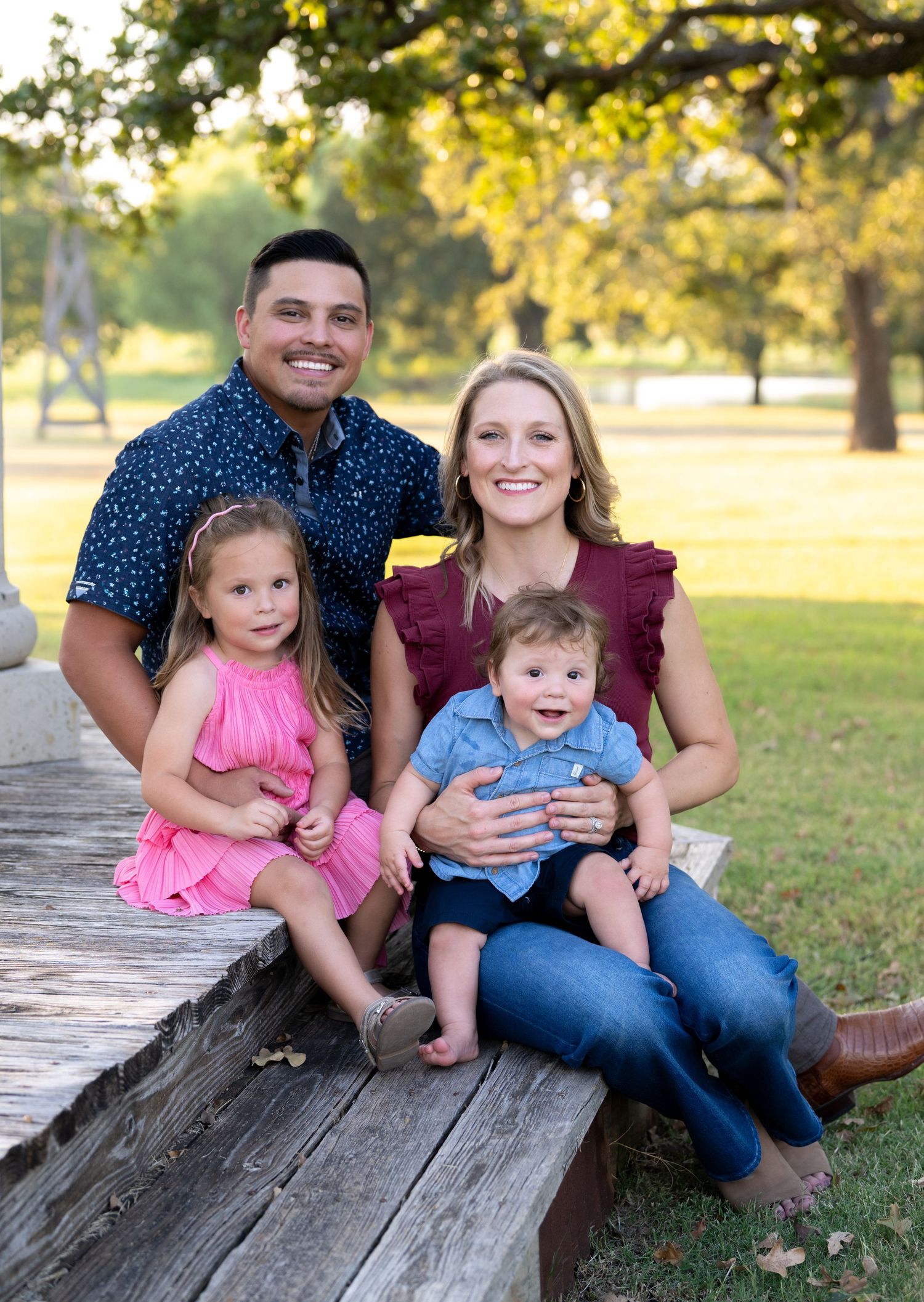 Family sitting together on rustic wooden bench in park during golden hour with autumn trees in background.