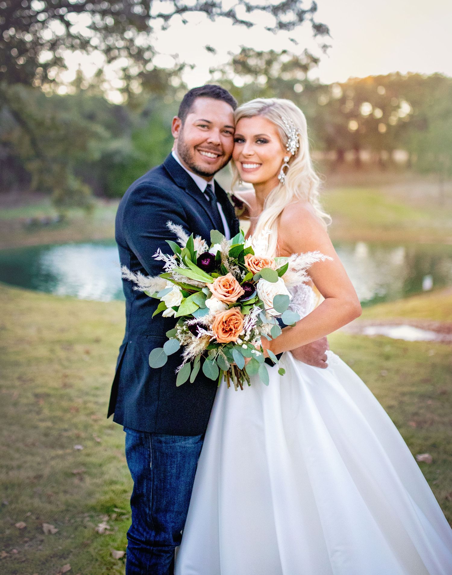 Newlyweds pose for a portrait at sunset near a lake with a colorful floral bouquet.