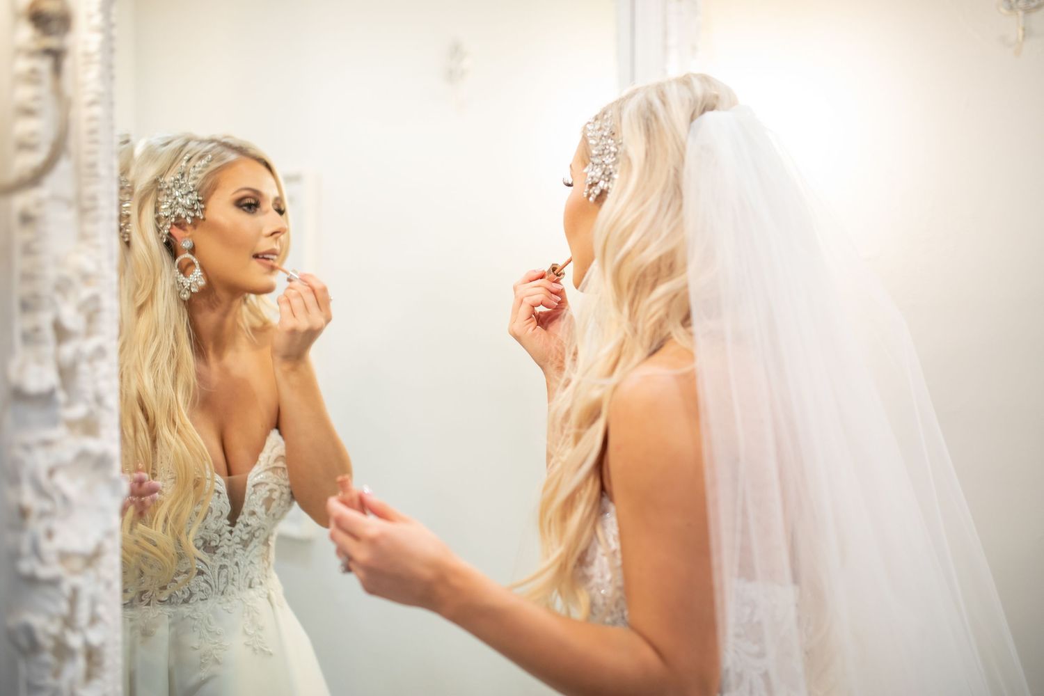A bride in a white wedding gown applies finishing touches while looking in an ornate mirror.