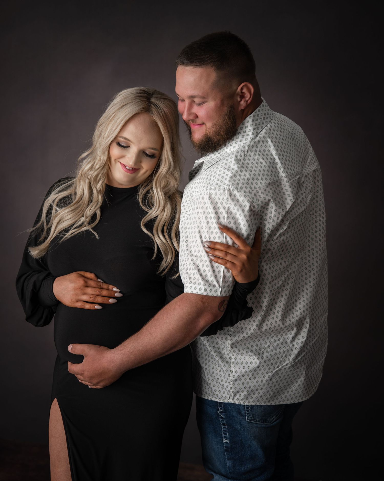 Intimate maternity photo session with couple against dark studio backdrop.
