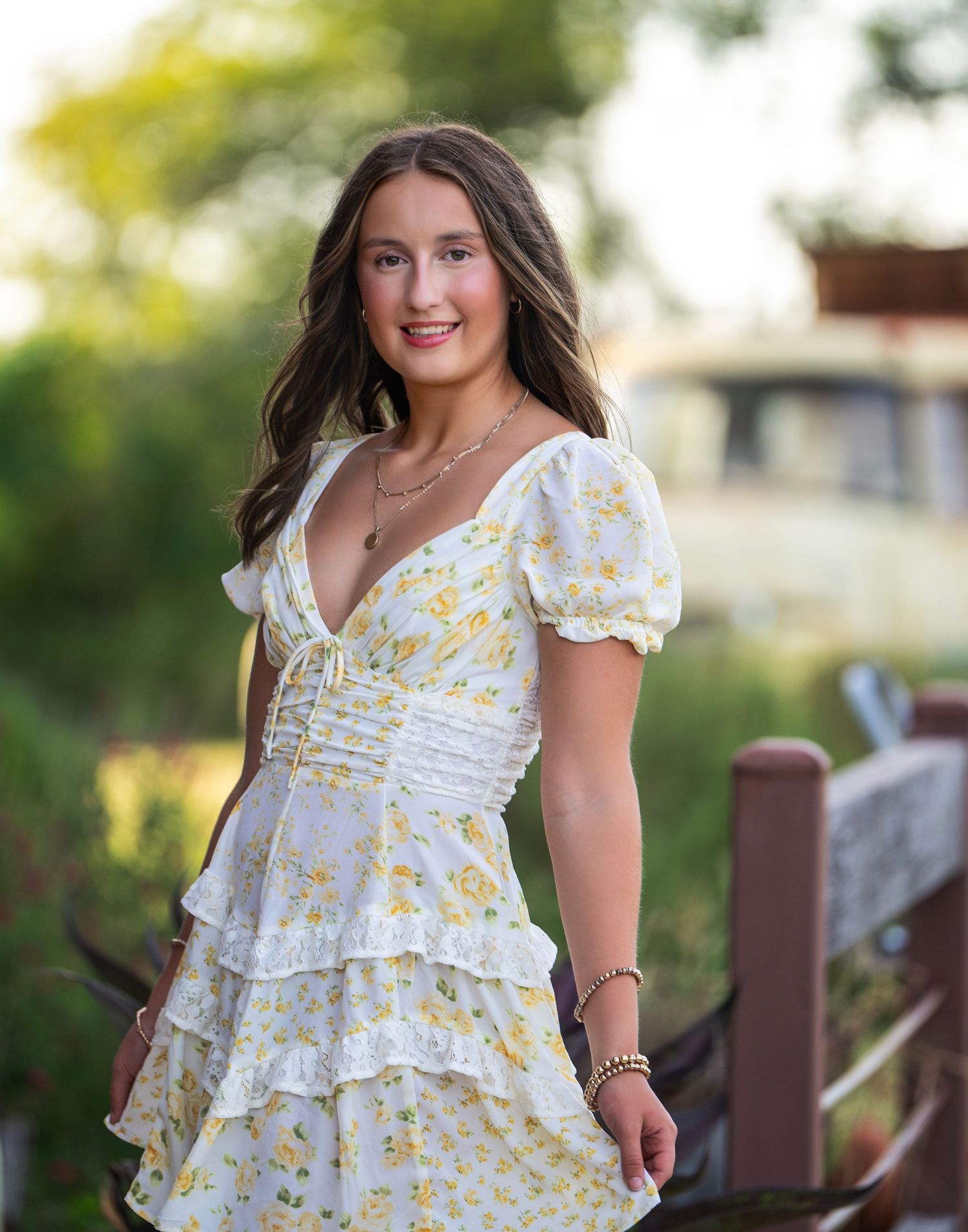 Young woman in a white and yellow floral print dress stands against a soft-focus outdoor background.