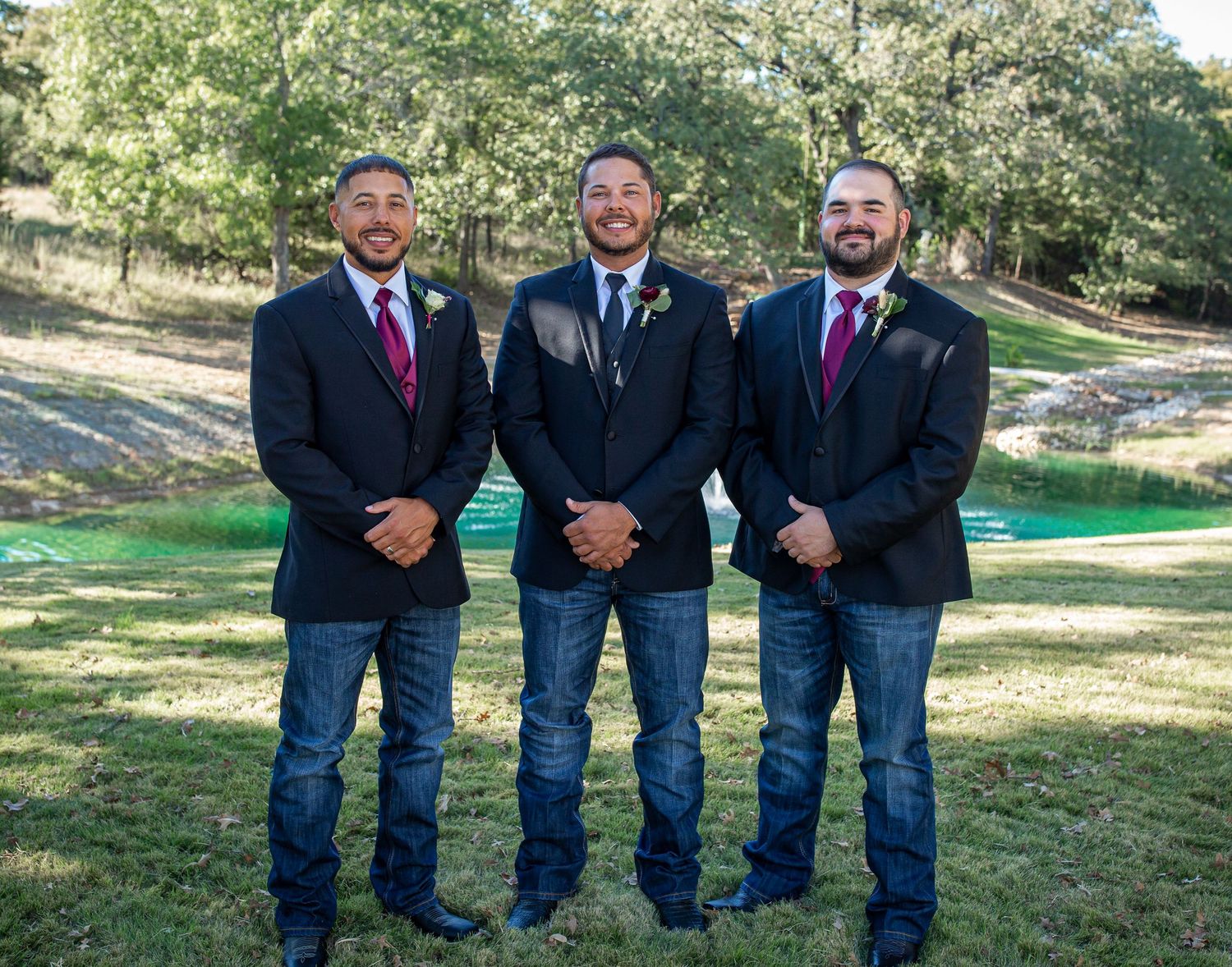 Three groomsmen in navy blazers and jeans pose together on grassy lawn.