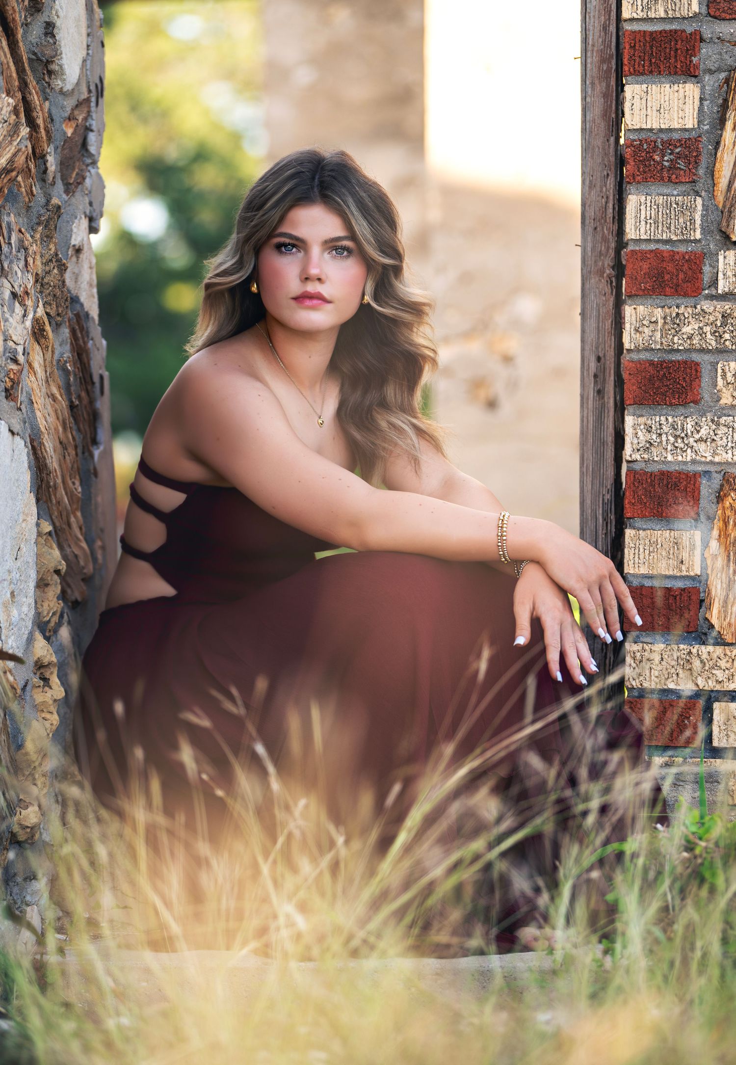 Artistic outdoor portrait session in tall grass with brick columns creating a dreamy golden hour atmosphere.
