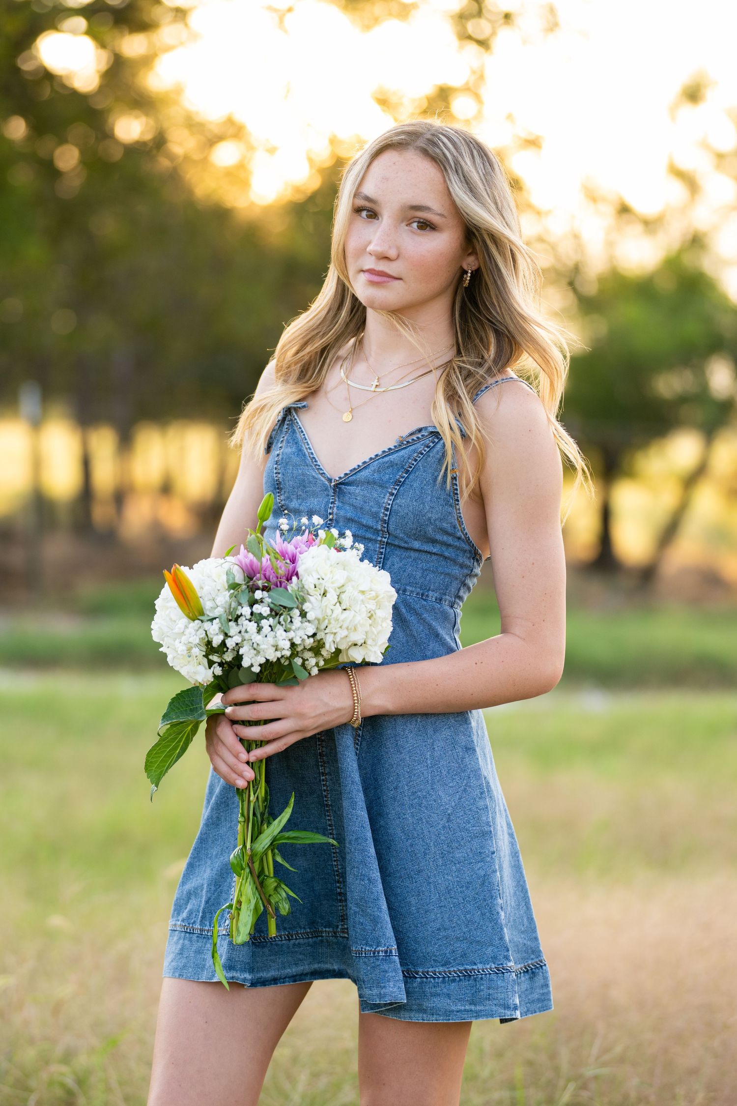 A person in a denim romper holds wildflowers while posing in a sun-lit field during golden hour.