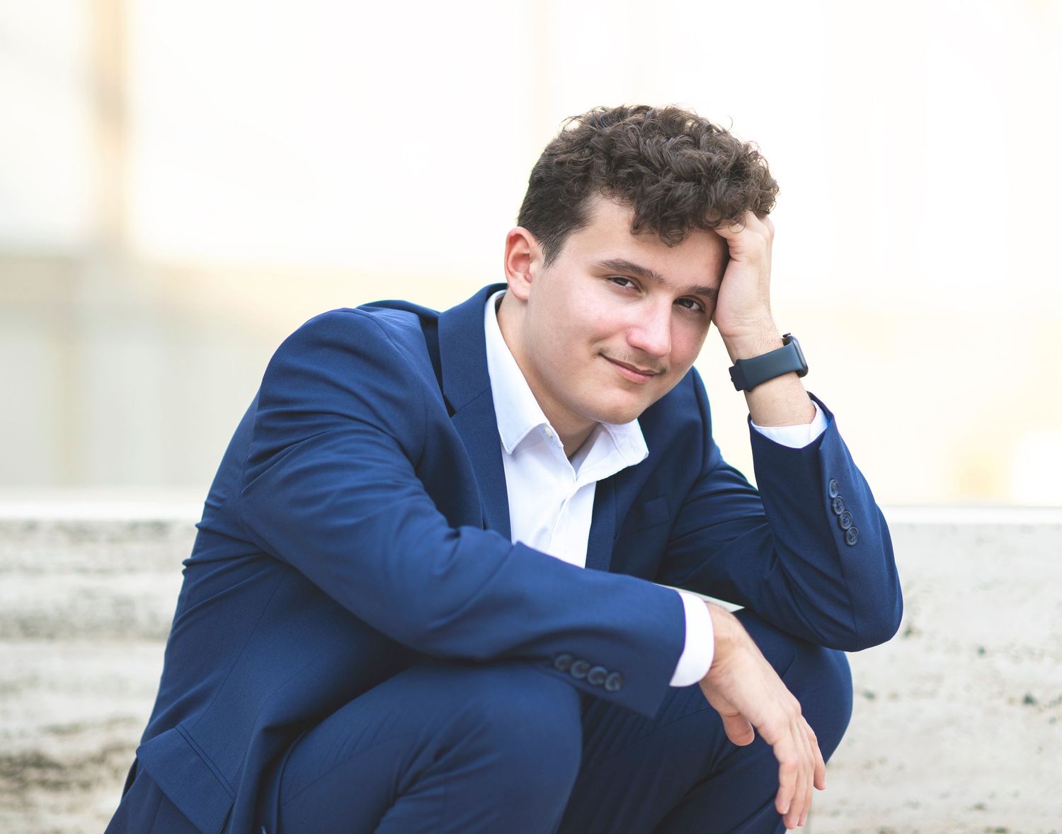 Young man in navy blue suit poses thoughtfully against neutral background.