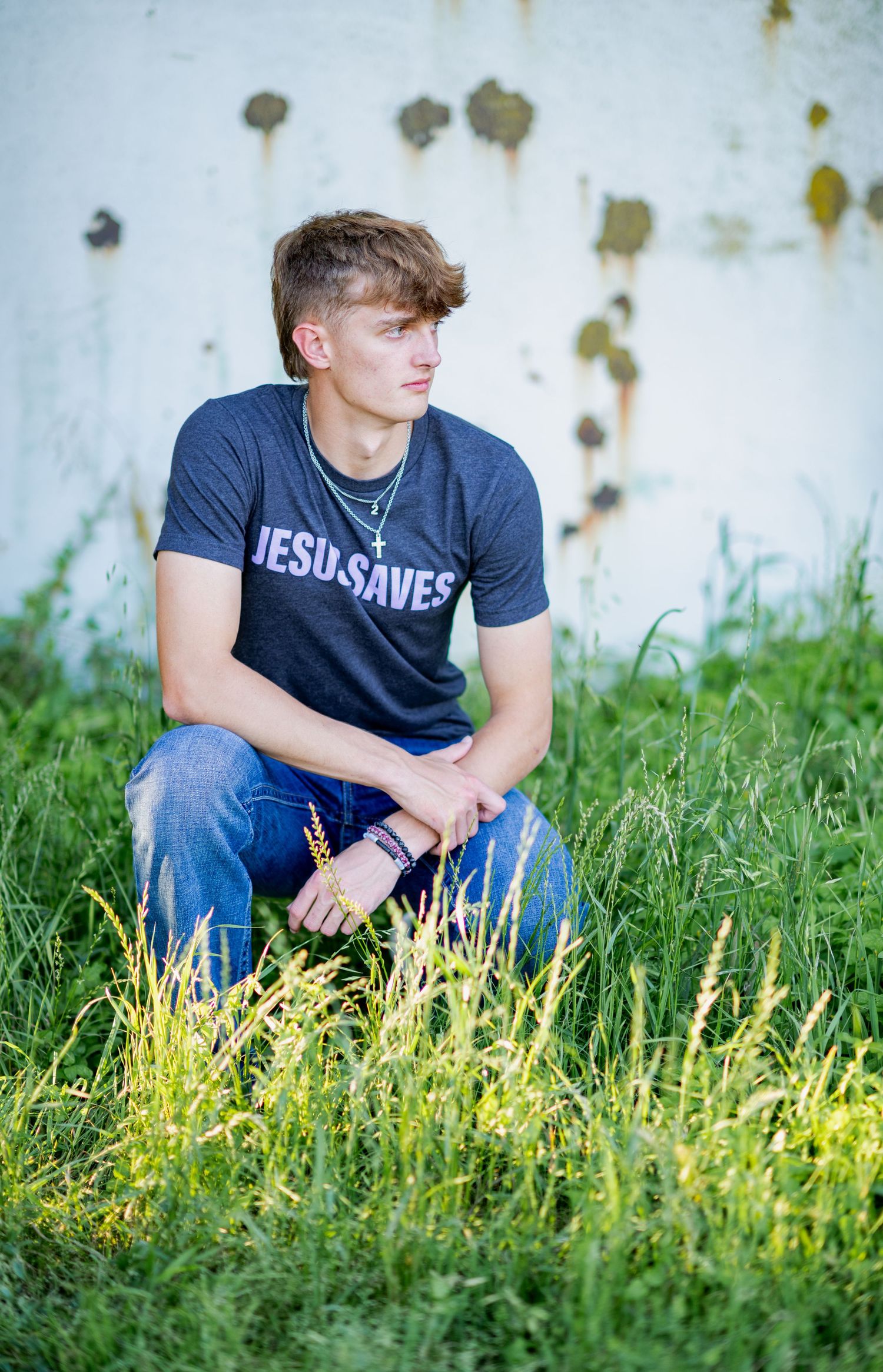 Person in navy Jesus Saves shirt kneels in tall grass for outdoor portrait.