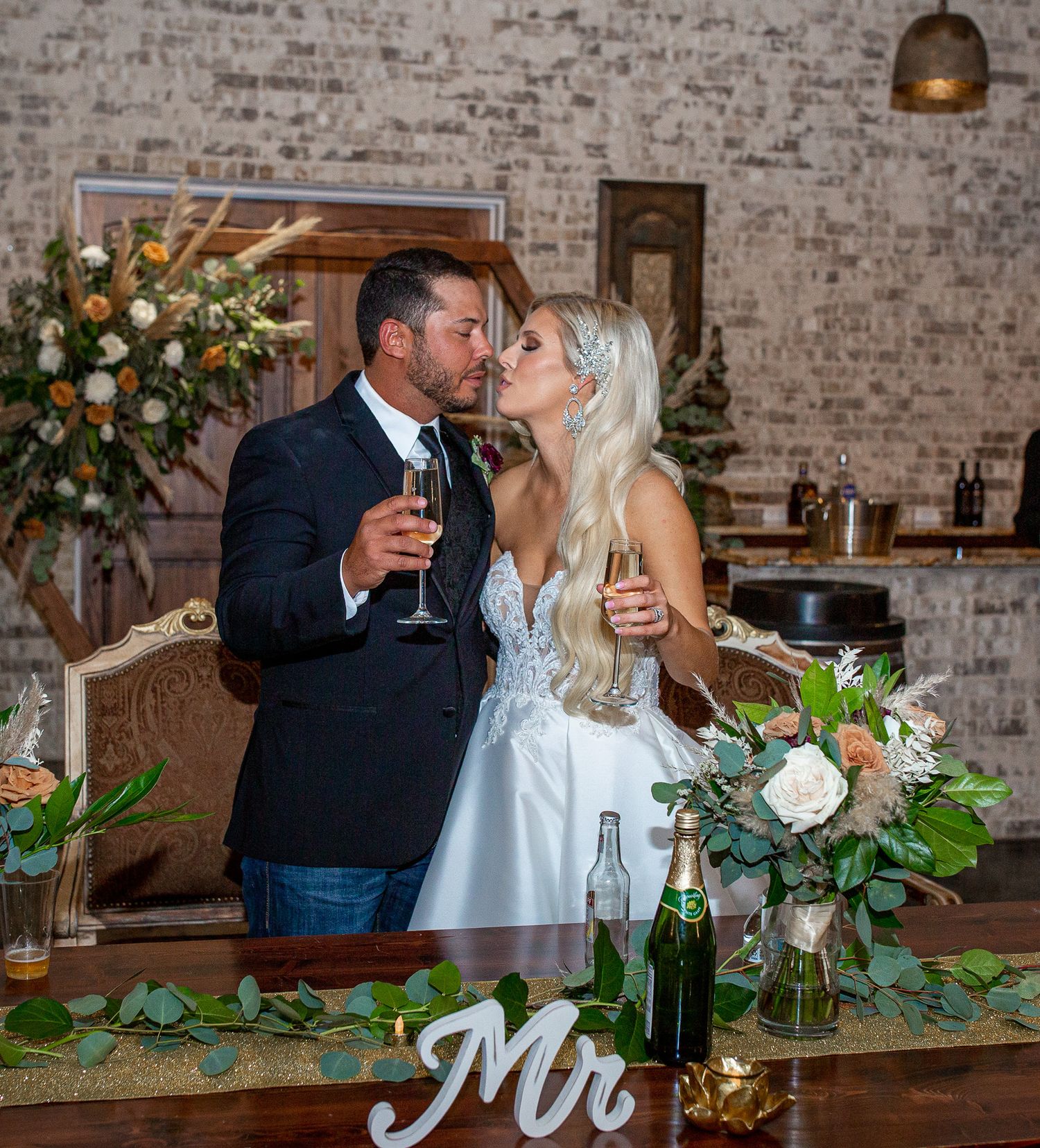 Elegant table setting at wedding reception with greenery garland and Mr sign decor against exposed brick wall.