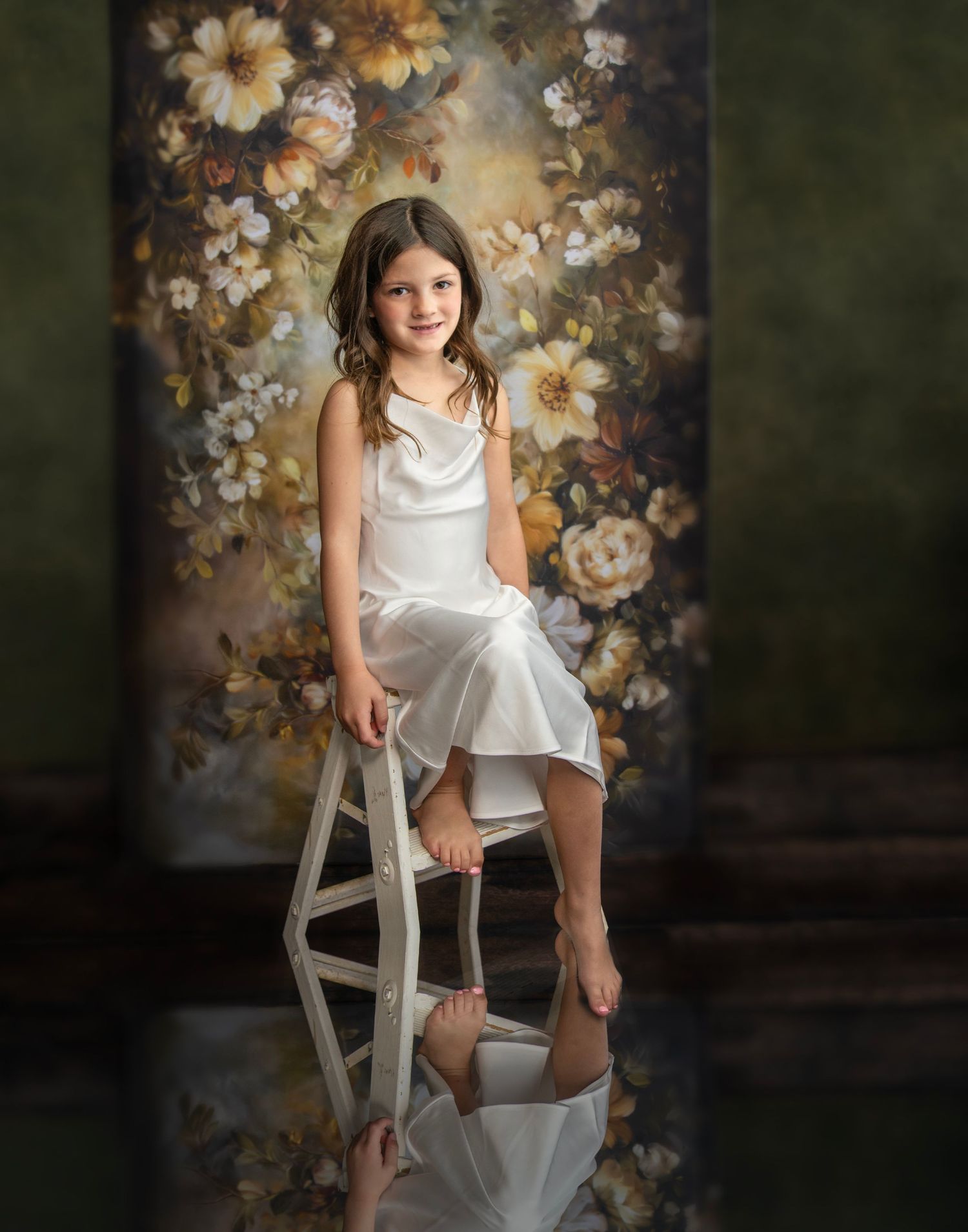 Child in white dress sits on wooden stool against floral backdrop in artistic portrait setting.