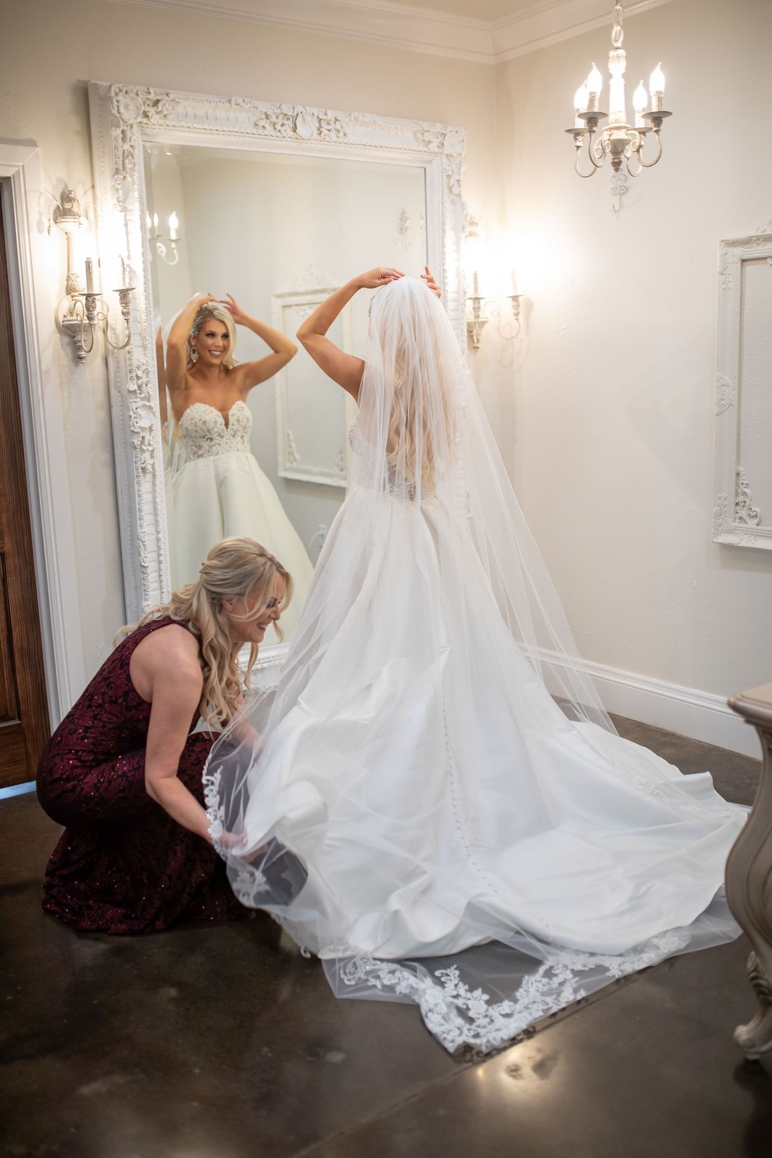 Wedding dress being arranged on floor in elegant dressing room with crystal chandeliers.