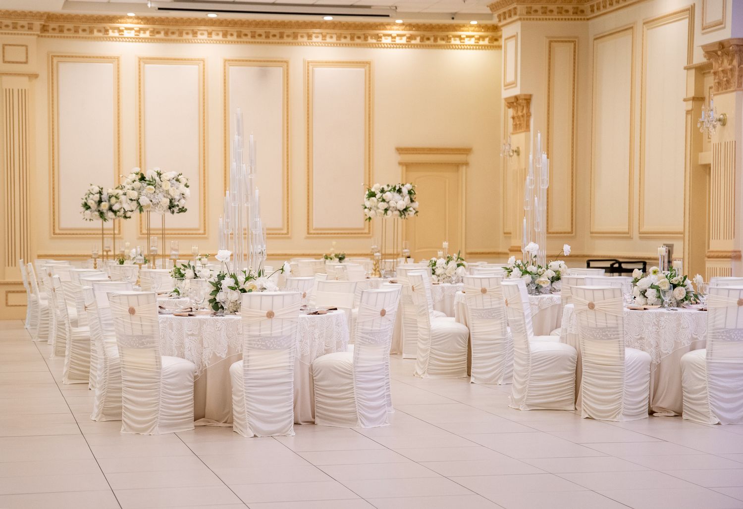 Elegant white and gold ballroom set for wedding reception with round tables and white chairs.