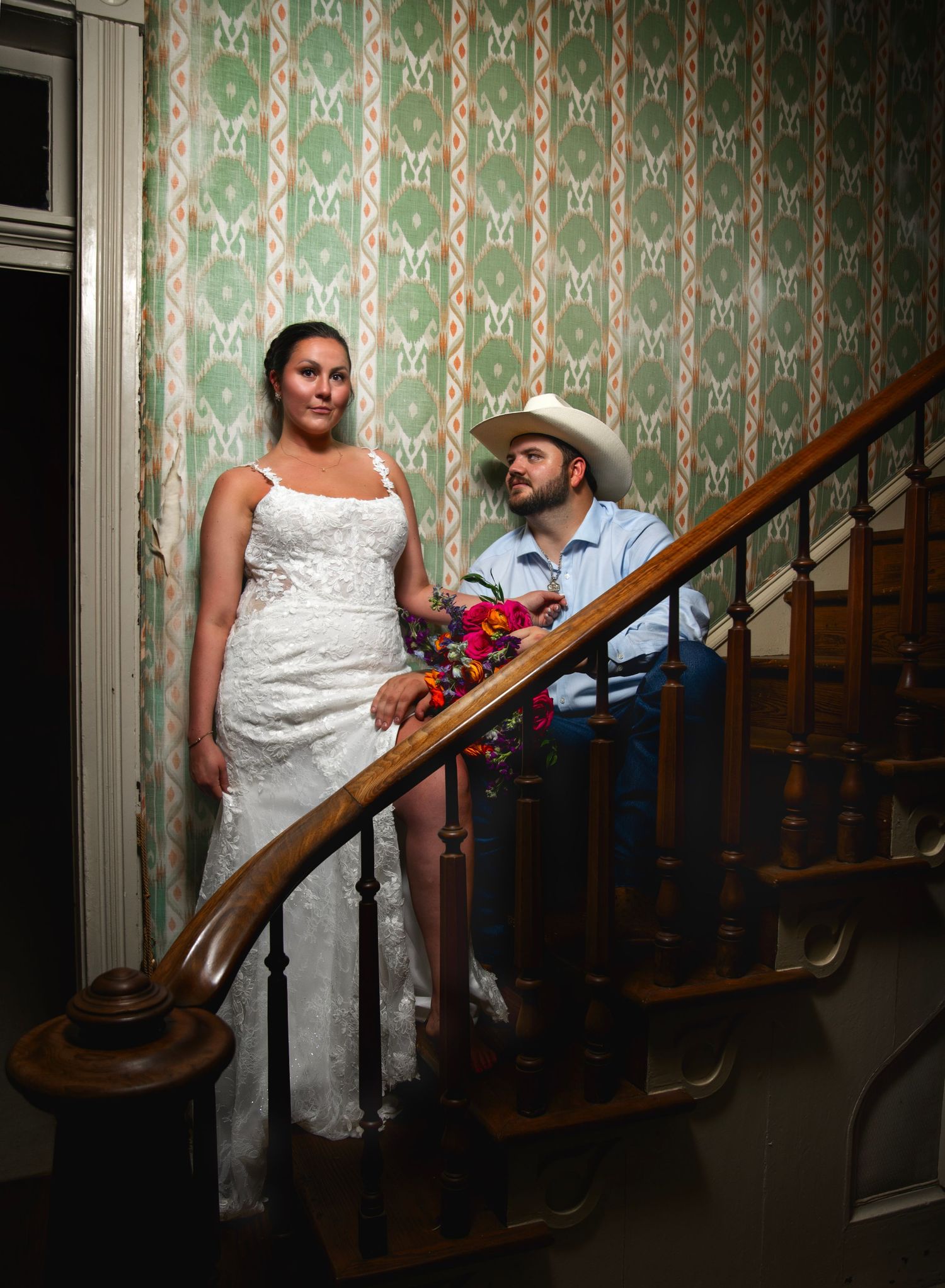 A vintage-inspired wedding photo on a staircase with green floral wallpaper.