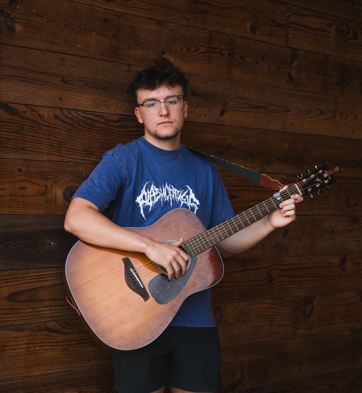 Person playing acoustic guitar against wooden wall background wearing blue t-shirt.