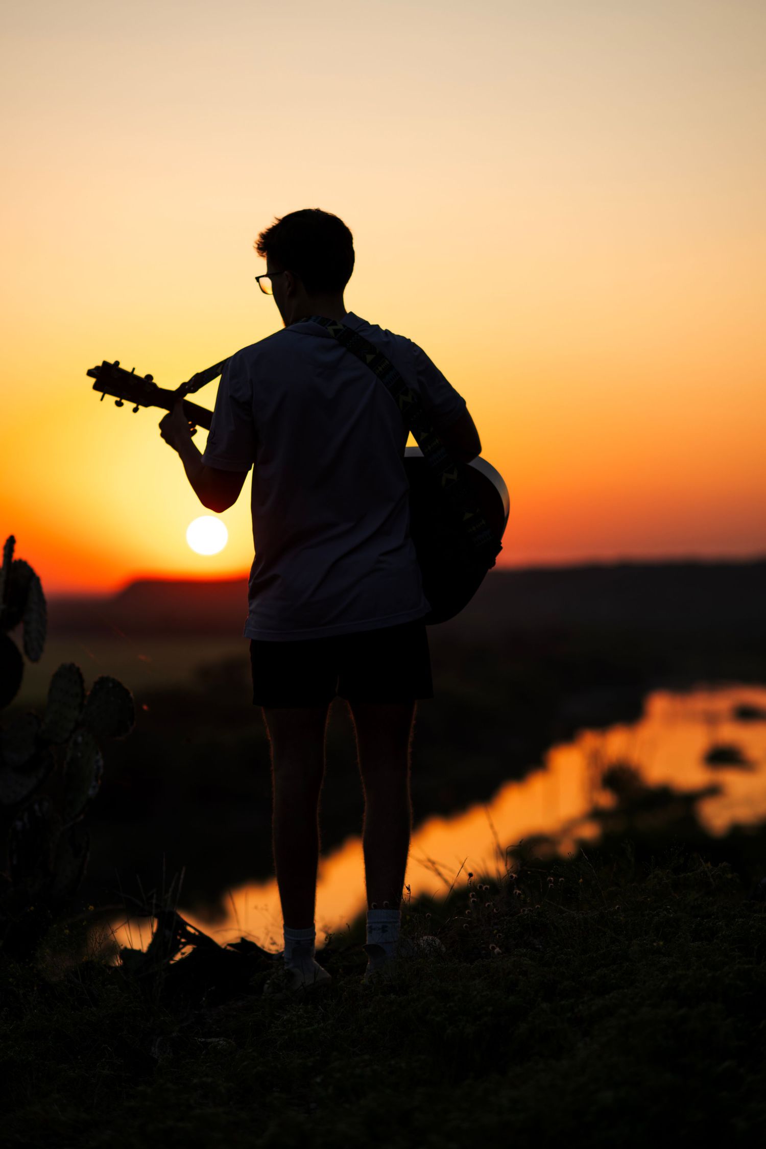 Silhouettes of musicians playing guitars at sunset by a lake.