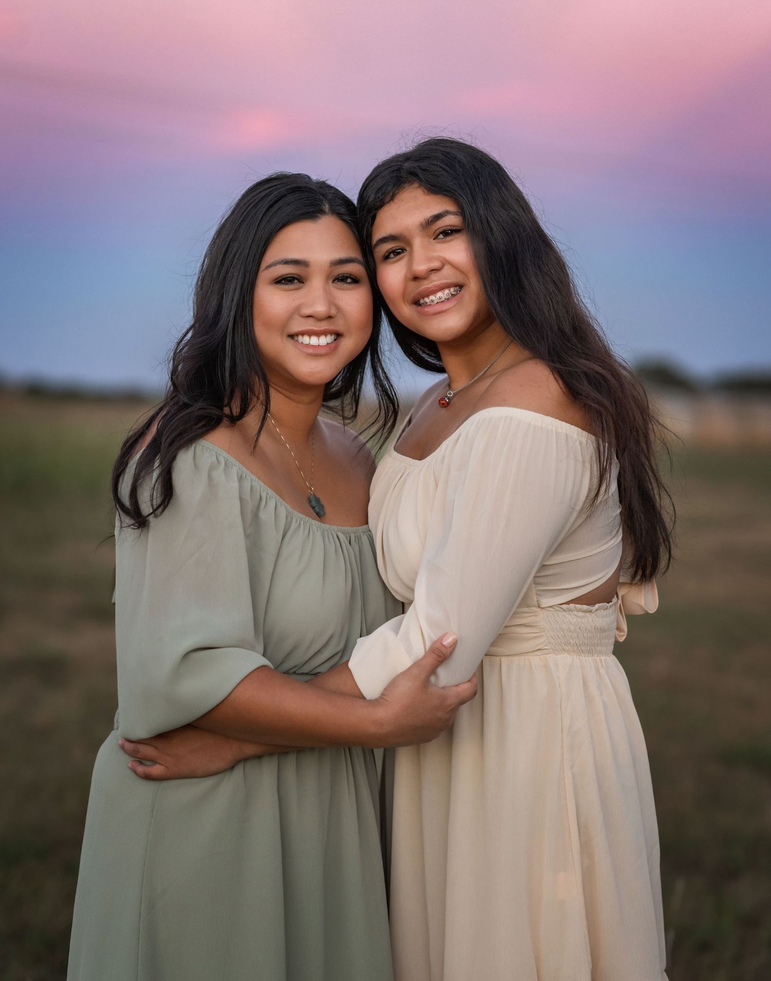 Two women in flowing dresses embrace against pink and blue sunset sky.