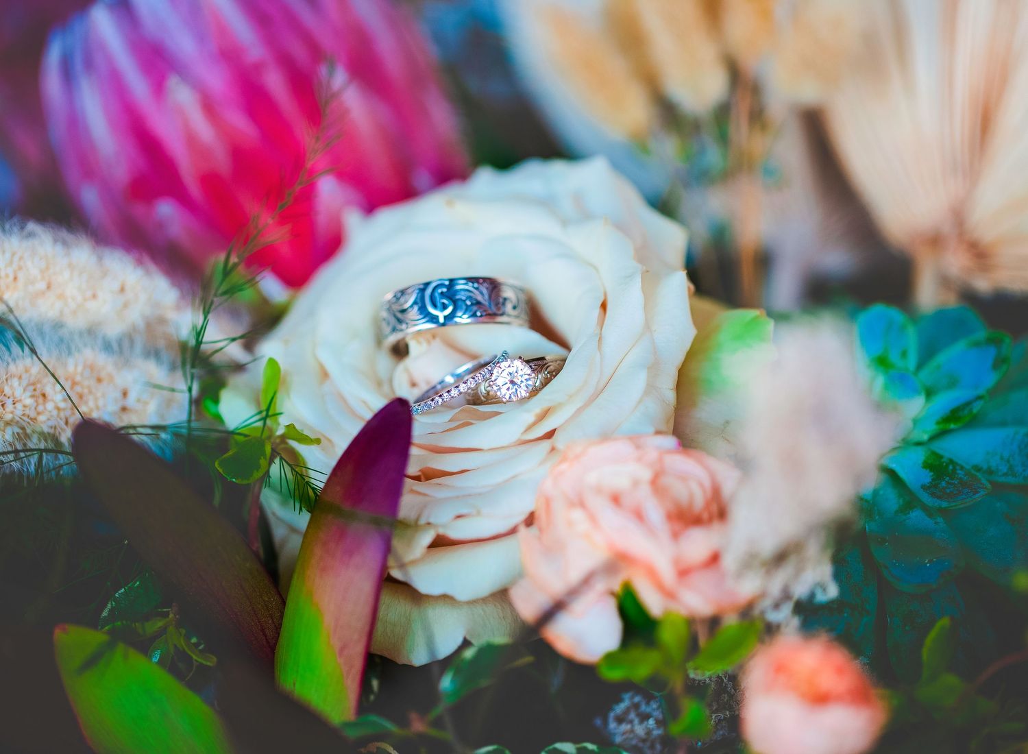 A white rose with wedding rings nestled in its petals, surrounded by colorful flowers in soft focus.