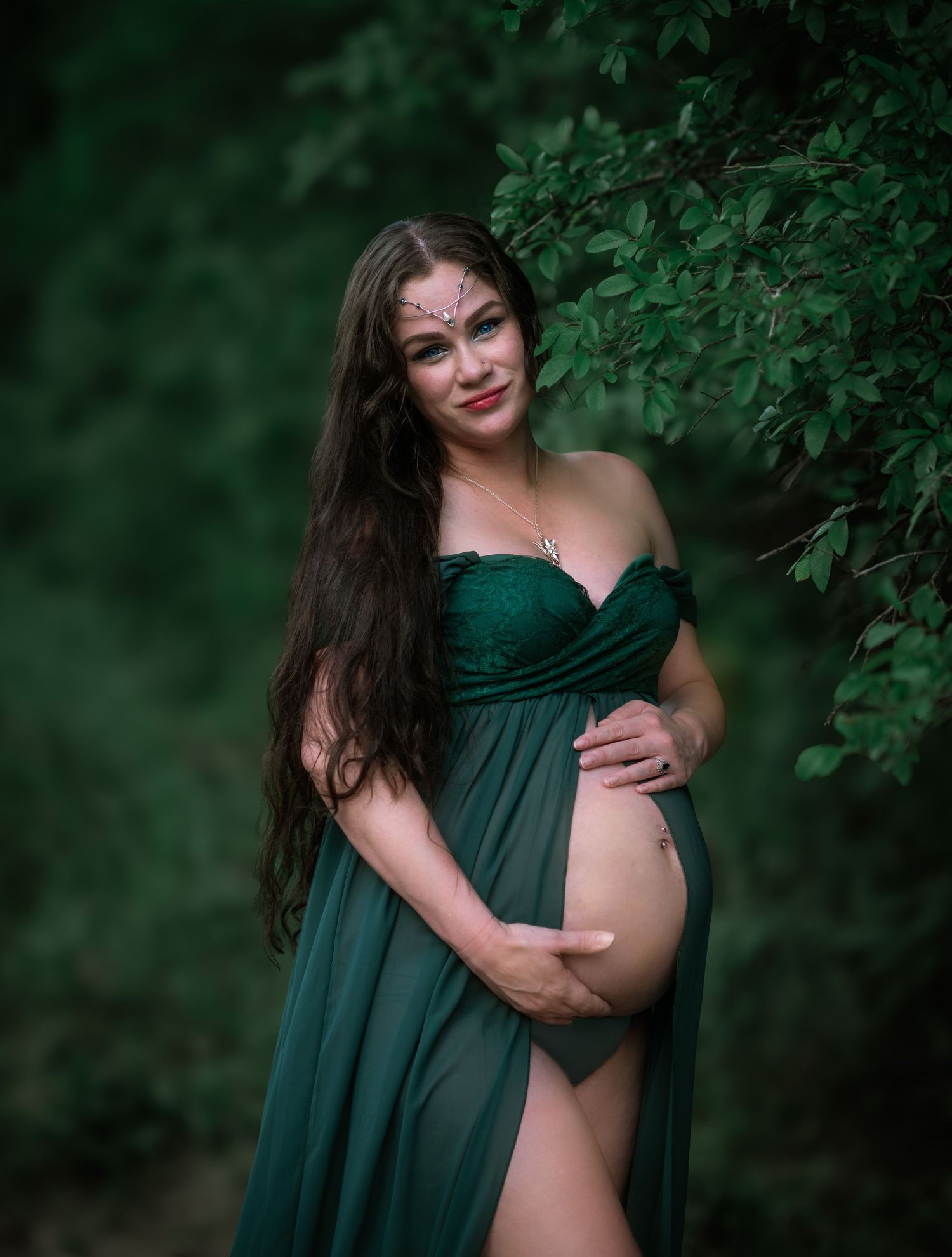 Natural maternity portrait amidst green foliage wearing emerald off-shoulder gown.